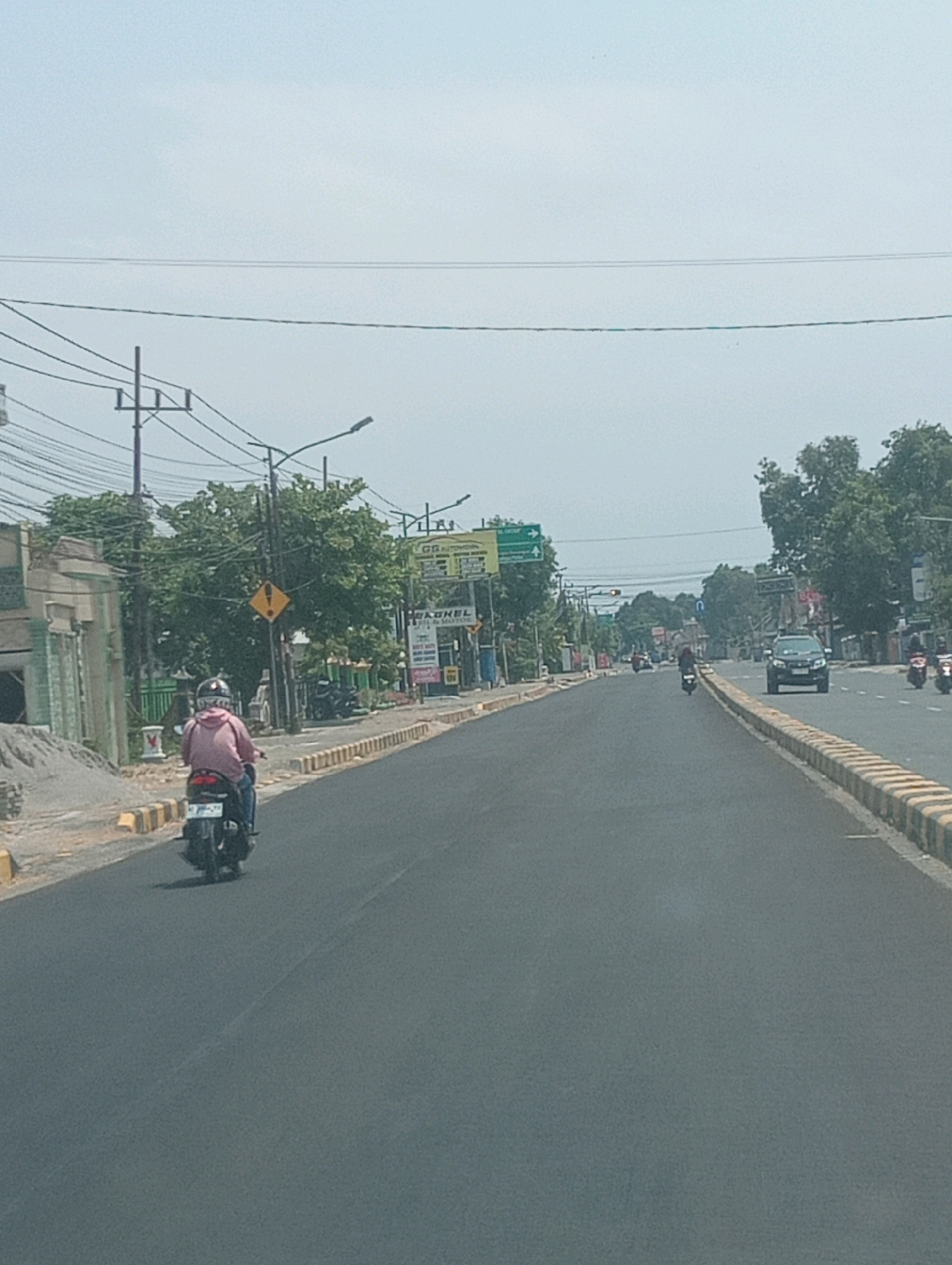 The image captures a street scene in Plosojenar, Indonesia, under an overcast sky. The perspective is from a vehicle looking forward, showing a two-lane asphalt road. On the left side of the road, there is a pile of sand and building materials, a light-colored building with a greenish trim, and a utility pole with numerous wires. Further down the left side, there are trees and a yellow diamond-shaped warning sign with a black exclamation mark. On the right side of the road, there is a concrete curb with alternating yellow and white stripes, followed by a row of trees.
A person wearing a pink jacket and a helmet is riding a black motorcycle in the left lane, moving away from the viewer. Further ahead in the same lane, a single motorcycle is visible. In the right lane, a black SUV is driving towards the viewer, and behind it, two more motorcycles are also moving towards the viewer. There are streetlights and traffic lights visible along the road, as well as several commercial signs. The overall environment suggests a typical urban or semi-urban street in Indonesia.