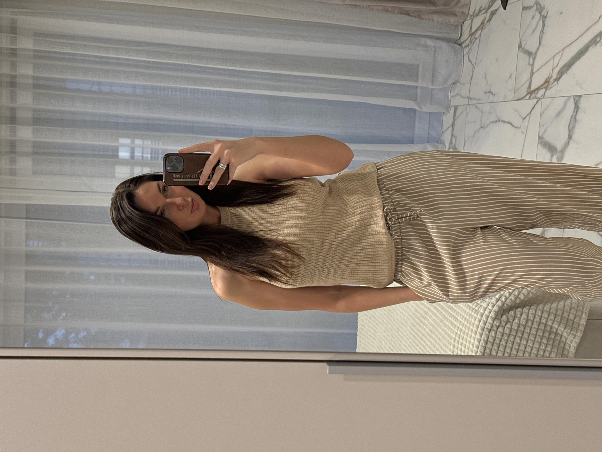 A young woman takes a mirror selfie. She is wearing a tan sleeveless top and tan and white striped pants. The background includes a window with white curtains, a marble wall, and part of a bed. The location is unspecified and there is no indication that it is Chaikovsky, Russia.