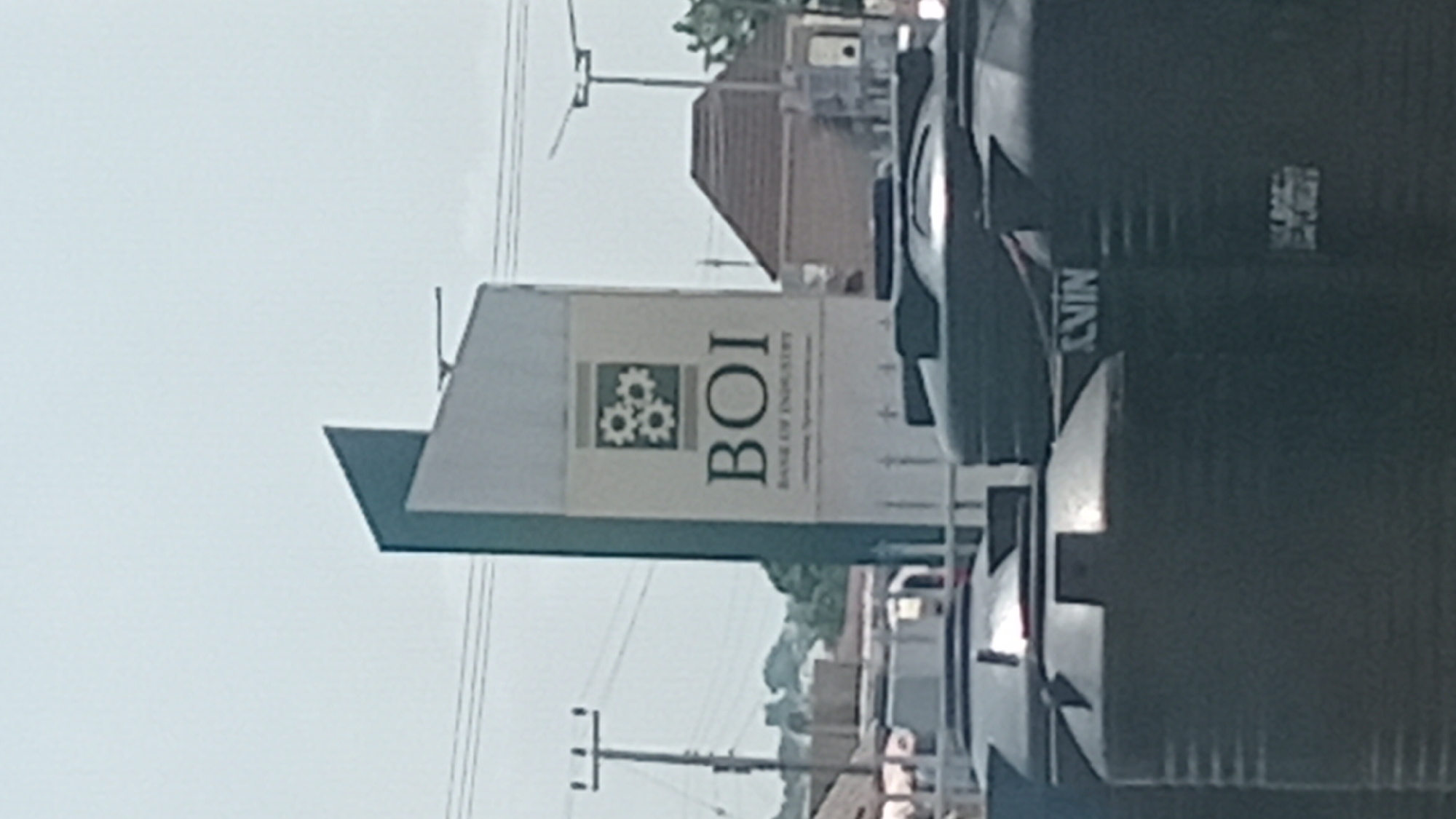 This image depicts a street scene in Jalingo, Nigeria, dominated by a prominent sign for the "BOI BANK OF INDUSTRY." The sign is tall and angled, with a green and white color scheme. At the top of the sign, there is a logo featuring three interlocking gears. Below the logo, the acronym "BOI" is displayed in large, bold letters, followed by the full name "BANK OF INDUSTRY" in smaller text. Further text, possibly a slogan or website, is visible at the bottom of the sign, but it is largely illegible due to the image resolution.
In the foreground and partially obscuring the lower part of the sign are several large, black water tanks. One of these tanks has the partially visible word "SVIN" printed on it in white. In the background, there are utility poles with electrical wires, a building with a brown roof, and some greenery, suggesting an urban or semi-urban environment. The sky is overcast, and the overall lighting suggests daytime. There are no people clearly visible in the frame.