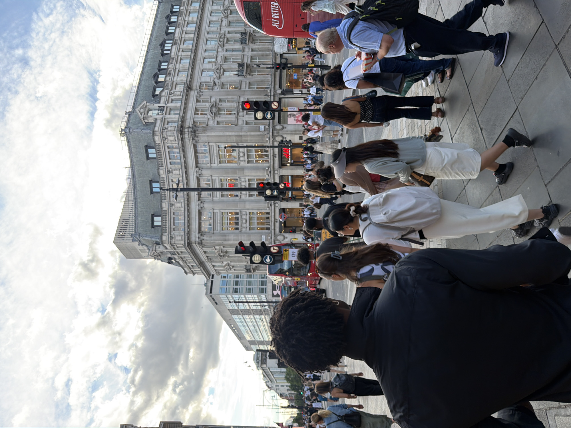 This is a street-level, eye-level, full-shot photograph taken in London, United Kingdom. The scene depicts a bustling street with pedestrians walking on the sidewalk. In the background, there are buildings, including a large, ornate building with a mansard roof and a modern building with a glass facade. A red double-decker bus, a common sight in London, is visible. Traffic lights are red. The sky is cloudy. People are dressed in casual clothing, suggesting it is daytime. The photograph captures a moment in time on a busy urban street.
