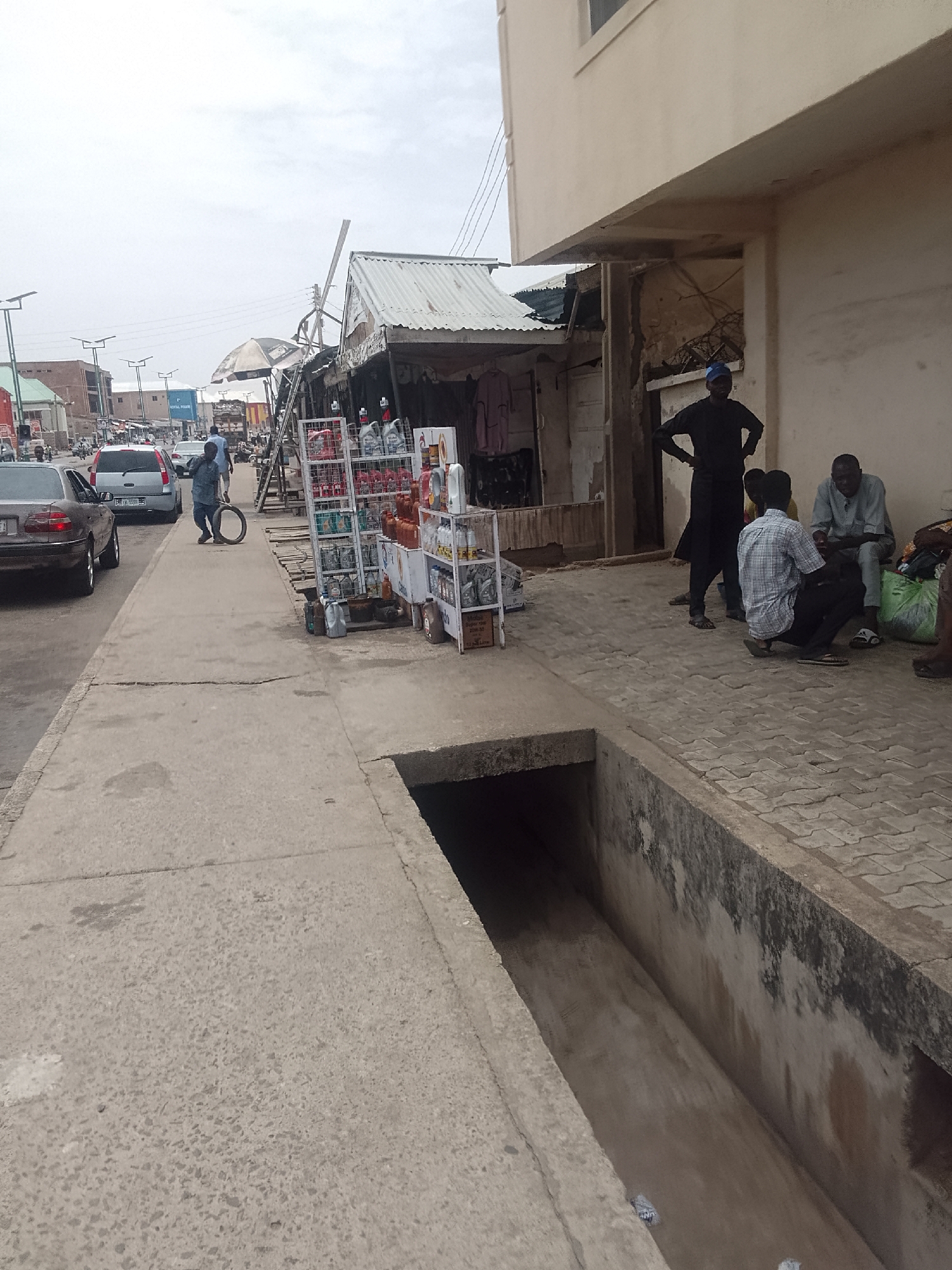 The image shows a street scene in Pantame, Nigeria. A drainage ditch runs along the foreground. A man is rolling a tire down the sidewalk. A makeshift kiosk displays motor oil and other automotive products. Nearby, a group of four men are gathered near a building, possibly conversing. There are cars in the background, indicating the scene is located on a street with vehicular traffic. The weather appears overcast. There are shops and buildings visible along the street.