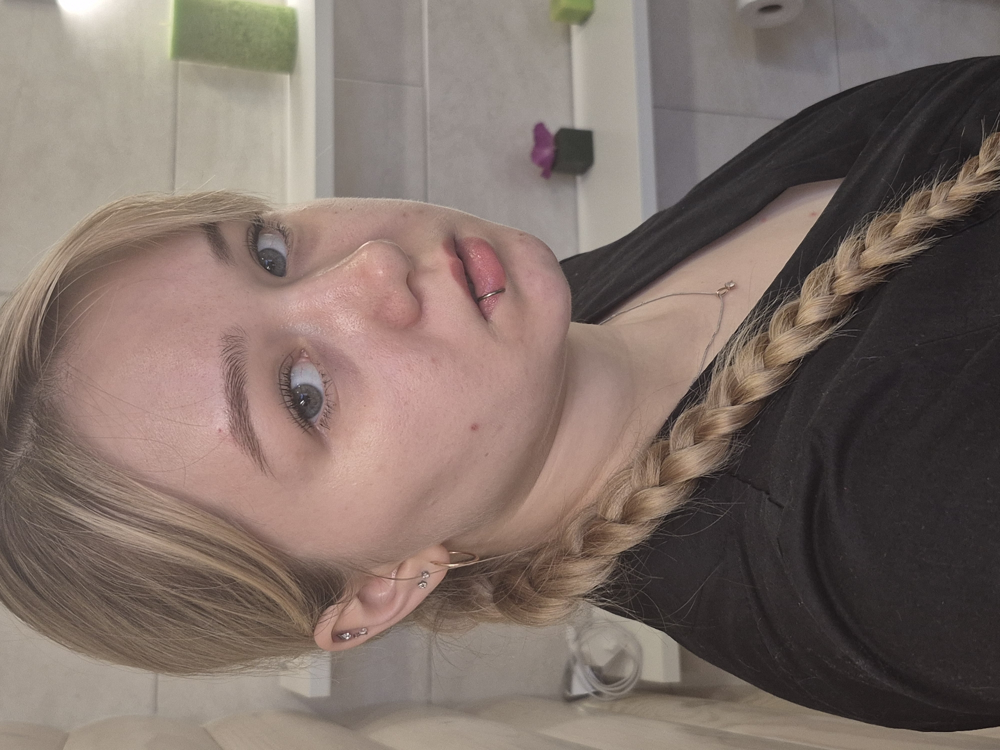 A close-up, vertically oriented photograph shows a fair-skinned young woman with light blonde hair, partially braided. She has blue eyes and a silver ring piercing in her lower lip. She is wearing a black top and a delicate silver necklace. Her left ear (as viewed by the observer) has multiple piercings and small earrings. The background appears to be a bathroom, with tiled walls and various objects including a green roll and a purple flower. There is no definitive information to conclude that the image was taken in Chaikovsky, Russia.