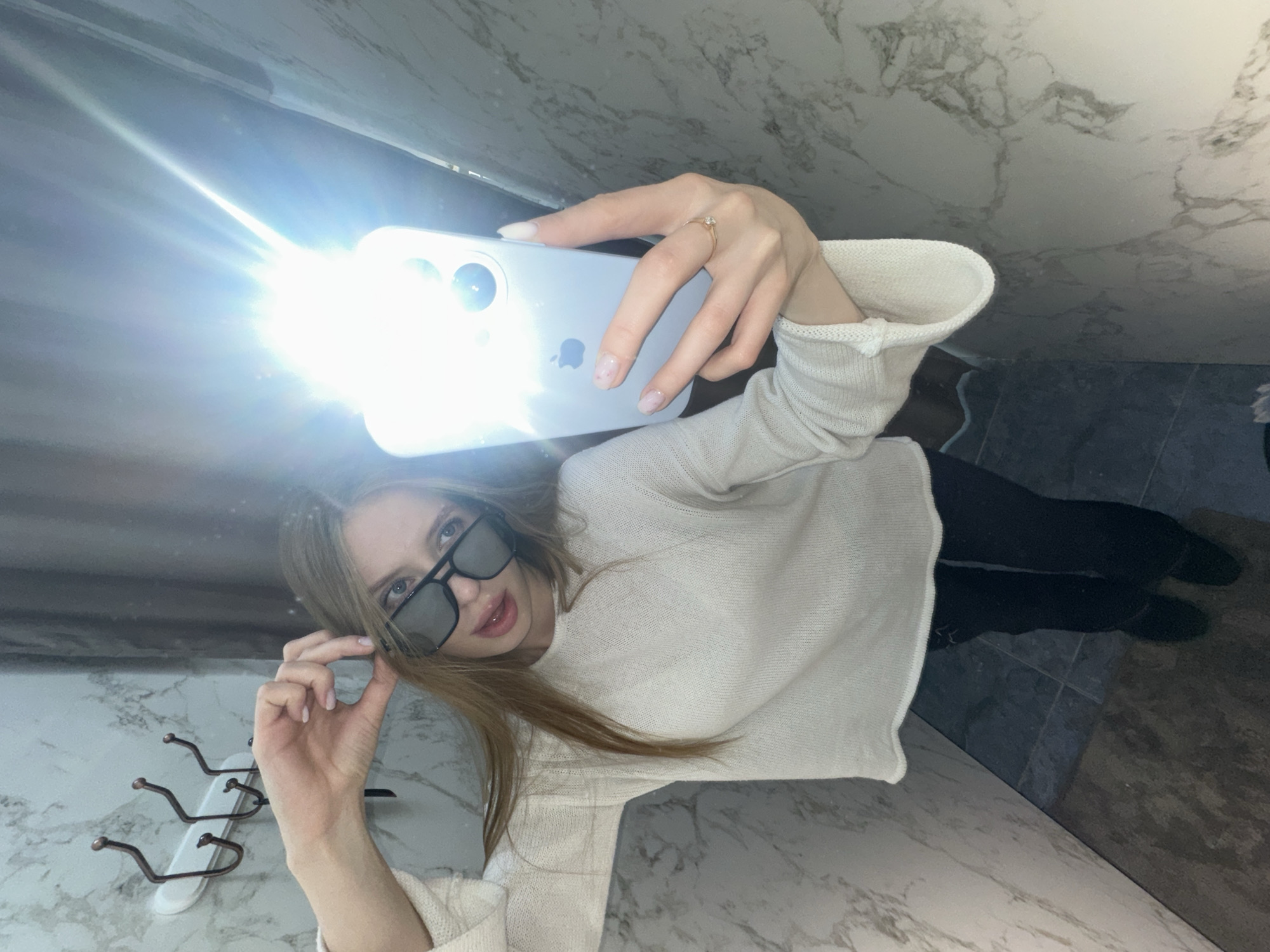 A woman is taking a selfie with a mobile phone with its flash on. She is wearing sunglasses and a white sweater. The location appears to be a bathroom based on the wall and floor tiles. A towel rack is visible on the wall. The location in Chaikovsky, Russia, cannot be verified from the image.