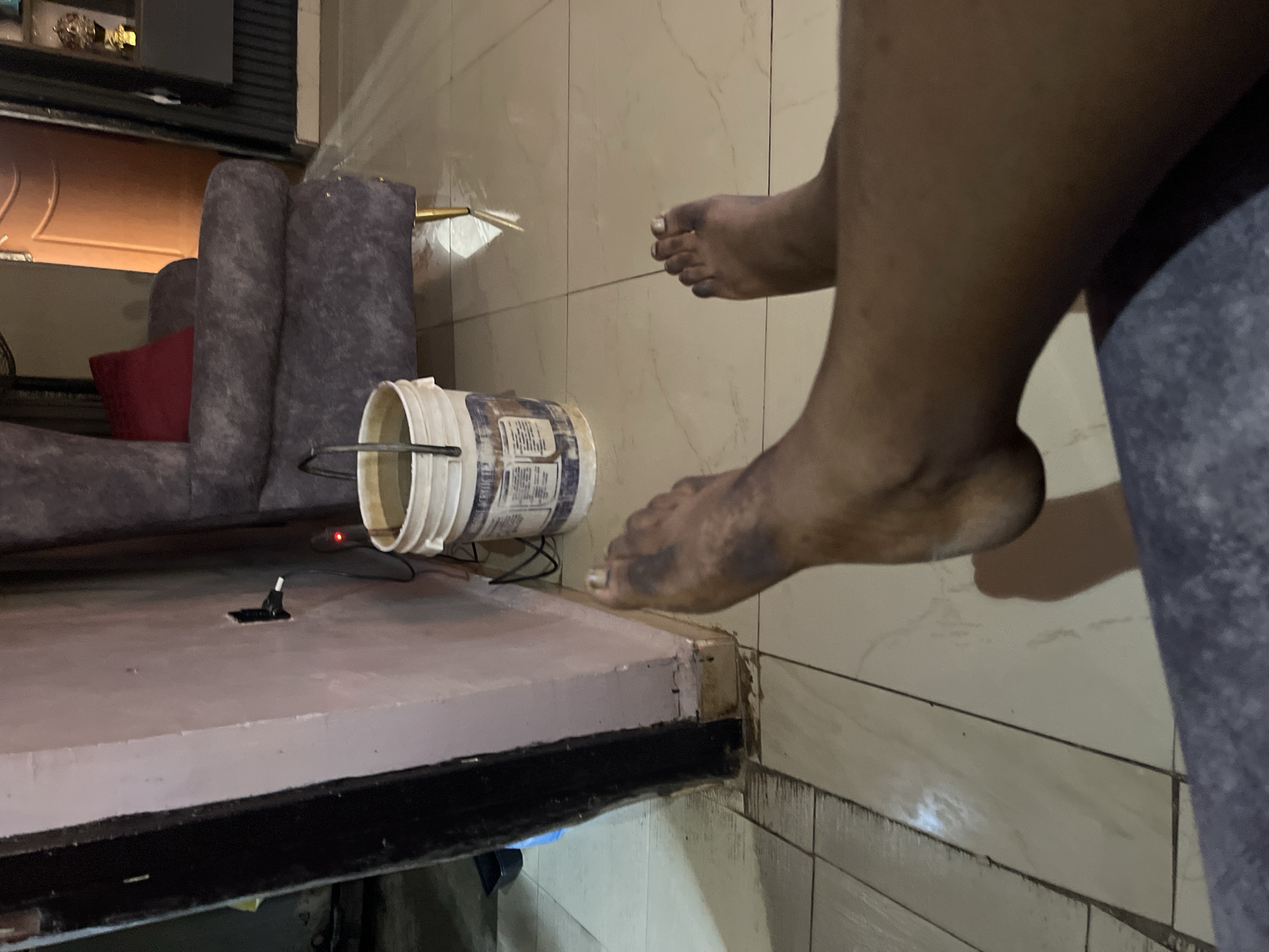 The image depicts a low-angle view of a person's bare feet on a tiled floor, suggesting they are sitting or lying down. The feet appear dirty, with dark smudges on the skin. In the background, a plush armchair with a red cushion is visible, along with a white bucket partially filled with a light-colored liquid, likely water. An electrical cord connects to a wall socket, and a small red light indicates a device is powered on. The environment suggests an indoor residential setting. The lighting is dim, with shadows cast on the floor. There are no specific visual cues that directly indicate the location in Jos, Nigeria, beyond the user's prompt to include it. The scene is static and focuses on the details of the feet and immediate surroundings.