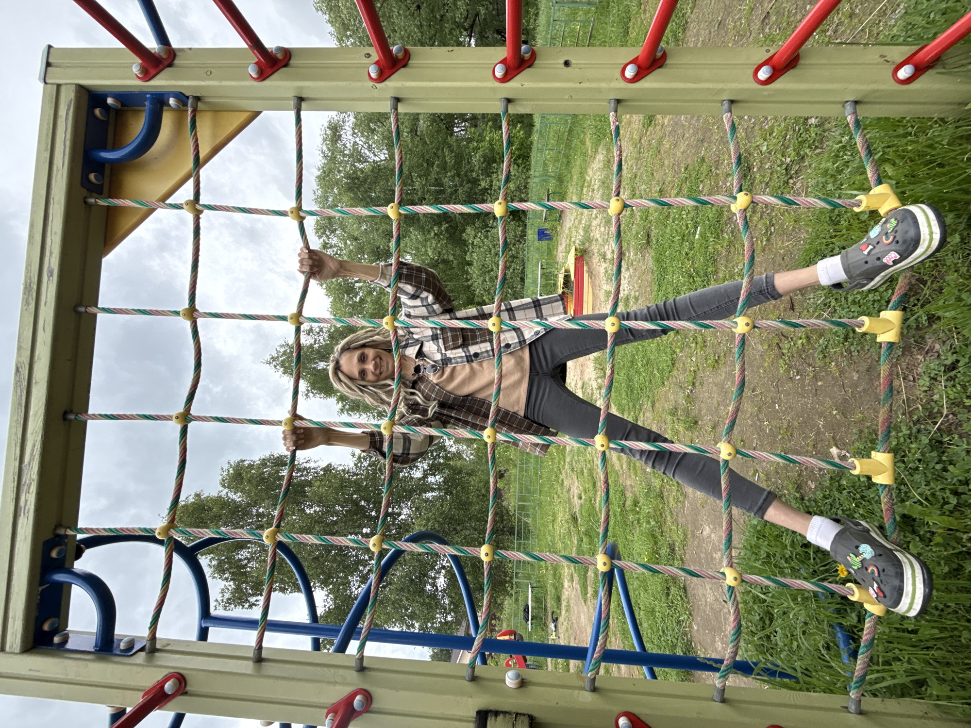 A woman is on a jungle gym in Rusanovo, Russia. The jungle gym consists of a green, red, and blue metal frame with a rope net. The woman is hanging onto the net by her hands and feet. She is wearing a brown and black plaid shirt, a tan shirt, gray pants, white socks, and black shoes. She has long blonde hair. The background shows a grassy area with trees and other playground equipment. The sky is overcast.