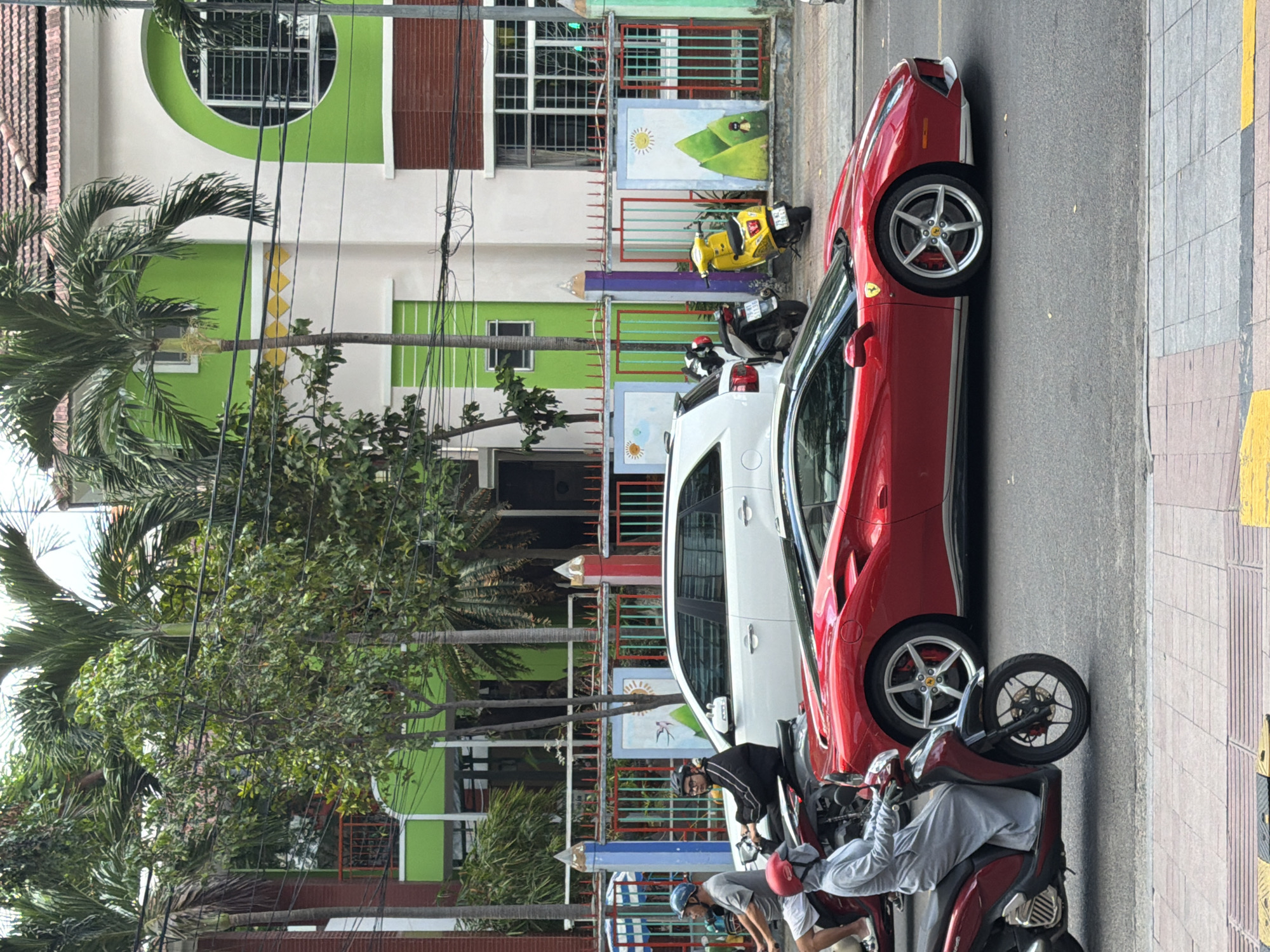 In Nha Trang, Vietnam, a vibrant street scene unfolds. A bright red Ferrari, a symbol of luxury, is captured in motion, its sleek design cutting through the urban landscape. In its wake, a white SUV and several motorcycles navigate the bustling road. The backdrop is a colorful building with green and white accents, adorned with playful murals of suns and mountains. Palm trees sway gently, their fronds casting shadows on the pavement. The scene is alive with activity, a snapshot of everyday life in this coastal city.