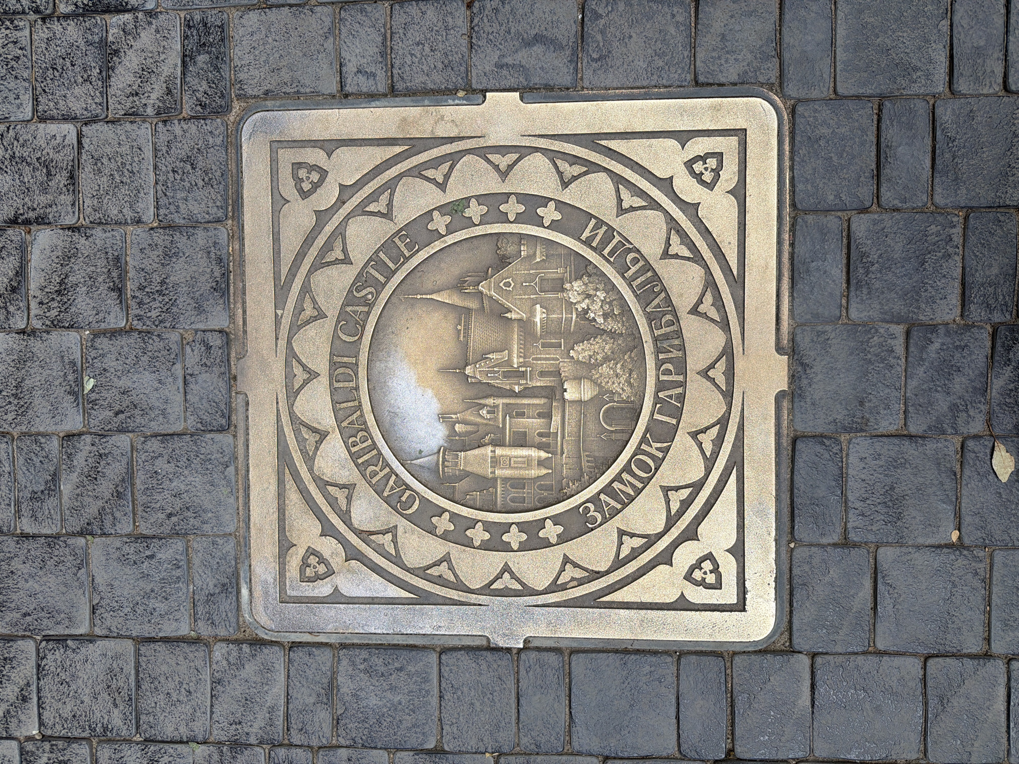 The image shows a square, bronze plaque embedded in dark gray, rectangular paving stones. The plaque features a circular design with an embossed image of a castle in the center. Surrounding the castle image are two concentric rings of text. The outer ring contains the English text "GARIBALDI CASTLE". The inner ring contains Cyrillic text that reads "ЗАМОК ГАРИБАЛЬДИ", which translates to "Garibaldi Castle". The plaque is worn and has a slightly tarnished appearance. The paving stones are arranged in a regular pattern and appear to be outdoors, possibly on a sidewalk or public square. There are no people or animals visible in the image. The lighting suggests it is daytime. The plaque is likely a historical marker or decorative element related to Garibaldi Castle in Khryashchevka, Russia.