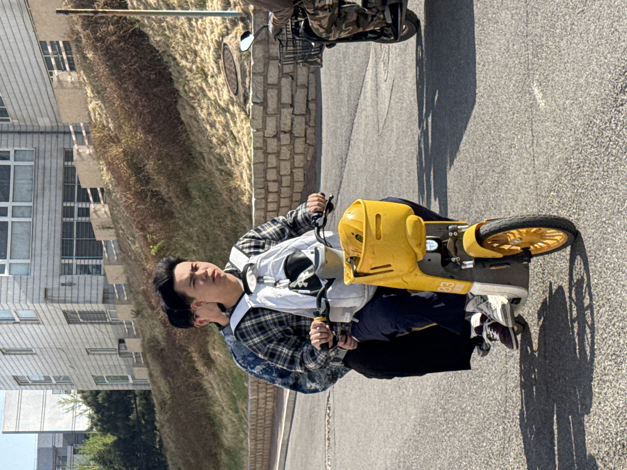 A bright, sunlit outdoor scene captures two individuals riding a yellow electric scooter on a paved road in Zhongtun, China. The image, taken during daytime, shows strong shadows cast on the asphalt, indicating clear weather.
In the foreground, a young man with dark hair is prominently seated on a yellow, three-wheeled electric scooter. He is wearing a plaid shirt and a white safety vest or harness. His hands are on the handlebars, and his gaze is directed slightly upwards and to his right. Partially visible behind him is another individual, likely also a young man, wearing a camouflage-patterned garment. The yellow scooter features a black seat and a yellow basket or container at the front, with "03" visible in black text on its side near the front wheel. The front wheel itself has yellow spokes.
To the left of the road, a sloped bank covered in dry, brown vegetation rises. Further left and in the background, several light-colored, multi-story buildings with numerous windows are visible, suggesting an urban or institutional setting. Green trees and bushes can also be seen further in the background.
The road itself is asphalt. A section of what appears to be a cobblestone path or barrier runs between the paved road and the grassy bank. A manhole cover is visible on this cobblestone area. In the upper background, another two-wheeled scooter or motorcycle, equipped with a rear basket, is partially visible, either parked or in motion. A vertical metal pole is also present on the far left of the upper background.