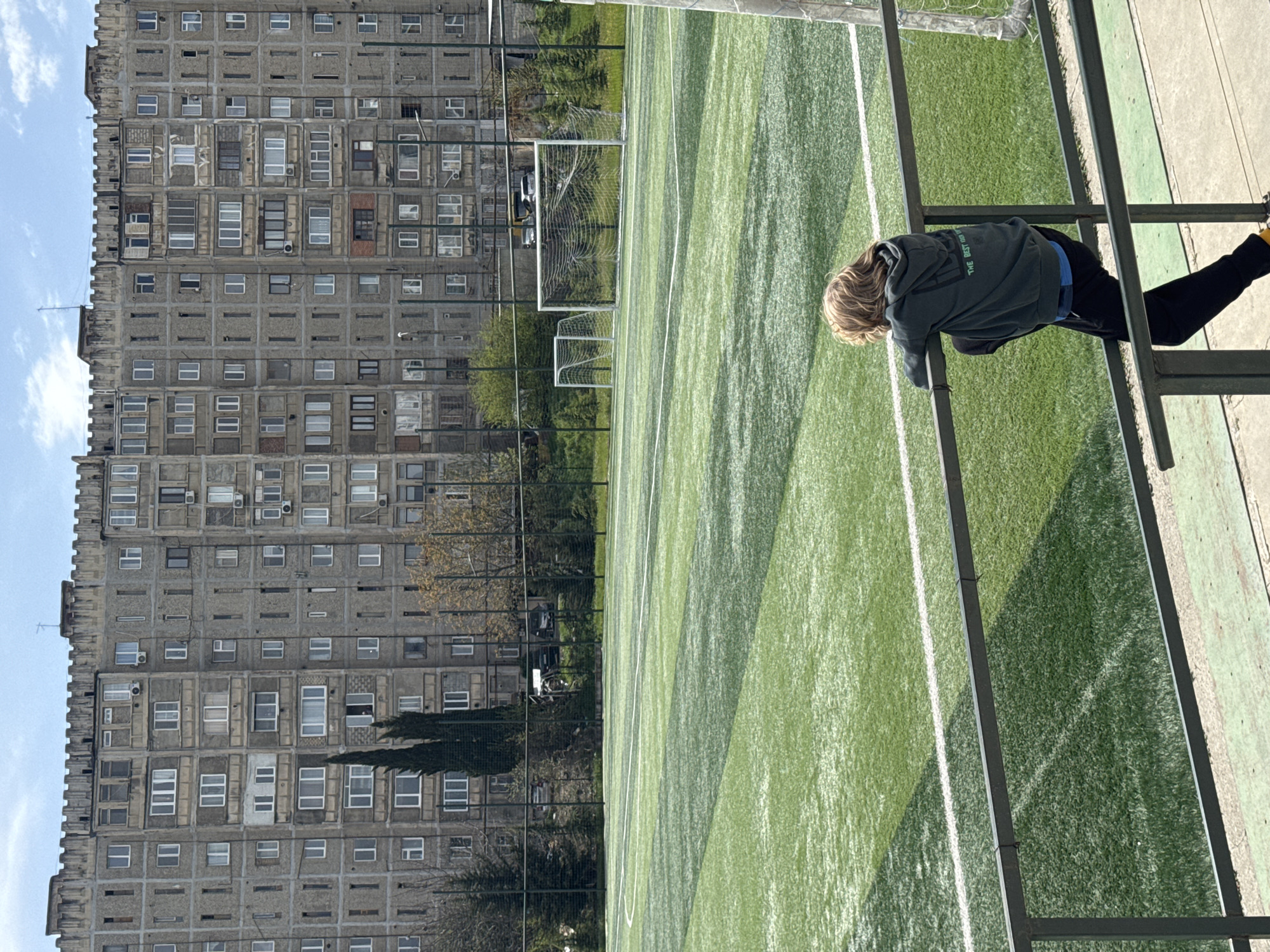 A high-angle, rotated shot reveals a person leaning on a metal structure, possibly a railing, overlooking an artificial turf soccer field. The person wears dark pants, a blue shirt, and a grey hoodie with lettering visible on the back. The soccer field has white lines marking its boundaries. In the background, a massive, concrete apartment building stands with rows of identical window openings. Behind the soccer field are trees and shrubs. The location is likely in Tbilisi, Georgia.