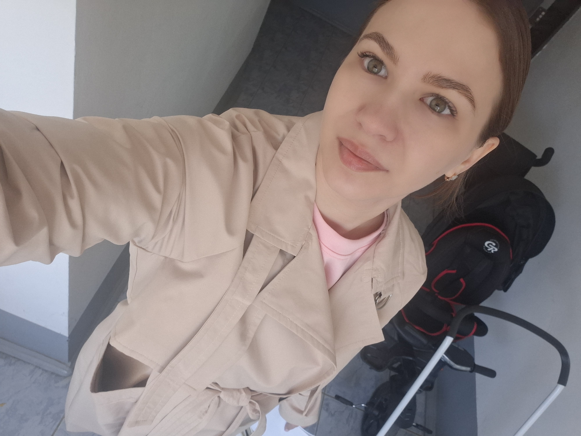 A woman wearing a beige trench coat over a pink top is posing for a selfie indoors. The background shows a gray tiled floor and a white wall, suggesting a hallway or entryway. A stroller is visible behind her to the right. The location is Chaikovsky, Russia.