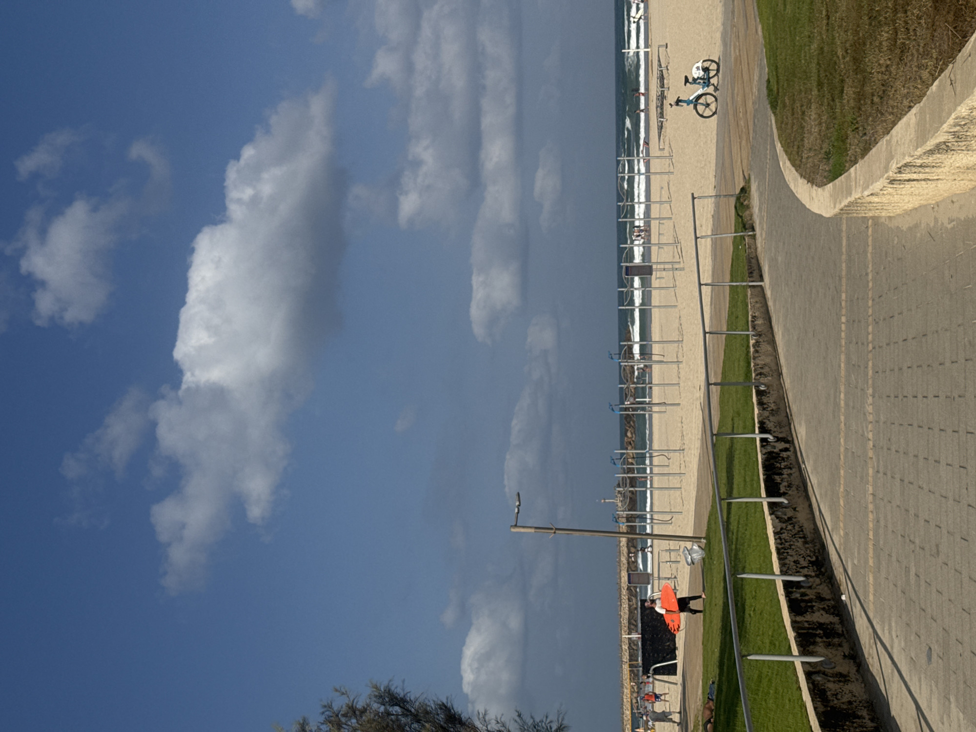 This image depicts a sunny day at a beach in Herzliya, Israel, characterized by a clear blue sky with scattered white clouds. In the foreground, a paved walkway curves along a grassy area bordered by a stone wall and a metal railing. Further back, the sandy beach is visible, with the Mediterranean Sea forming the horizon.
On the beach, there are several people engaged in various activities. A person in an orange shirt and black pants is walking with a bright orange surfboard. To the right, a blue and white bicycle is parked on the sand. In the background, a line of metal structures, possibly part of an outdoor gym or playground, stands against the sea. A jetty or pier extends into the water, with a lighthouse visible in the distance. The presence of surfers indicates active water sports. The overall atmosphere is that of a typical beach scene, popular for recreation and relaxation.