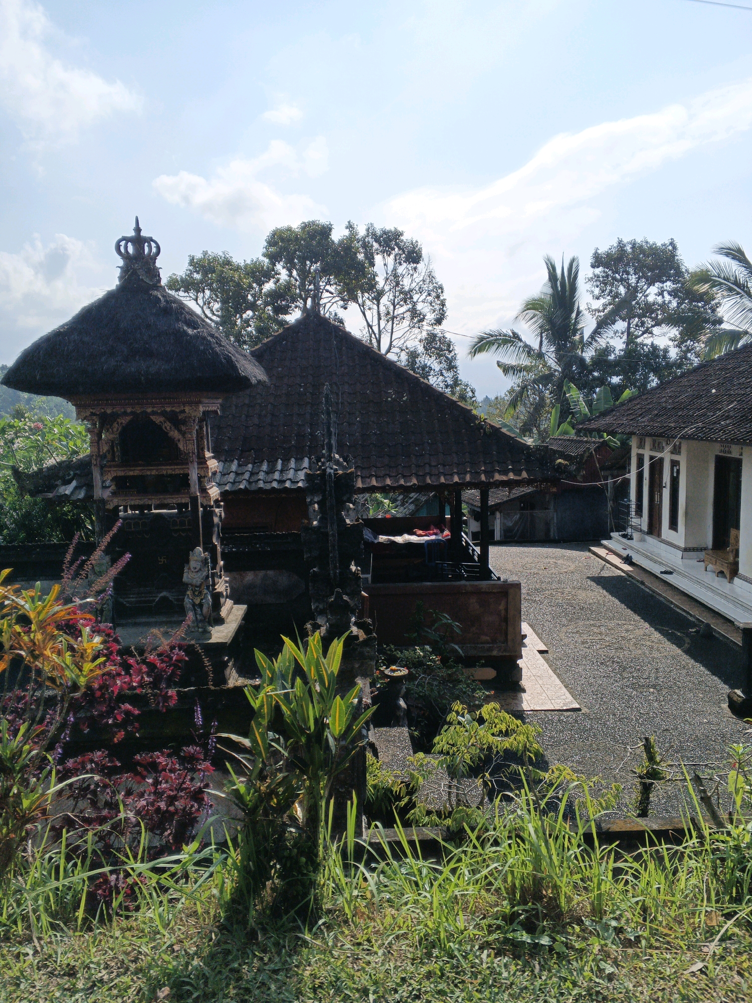 The image depicts a Balinese temple complex, likely in the Penebel region of Indonesia, bathed in bright daylight under a partly cloudy sky. The foreground is dominated by lush green grass and ornamental plants, including some with deep red foliage, with their shadows cast across the ground.
The focal point is a traditional Balinese structure with a multi-tiered, dark thatched roof, intricately carved wooden accents, and a decorative tiered finial resembling a crown. Adjacent to this is another building with a dark, tiled roof, partially obscured by trees. To the right, a white building with a dark tiled roof and dark framed windows and doors stands on a raised platform. The ground in the courtyard is covered with dark gravel.
The architectural style suggests a Hindu temple, common in Bali. The presence of statues and symbolic carvings, such as a swastika, further indicate its religious nature. The surrounding environment is verdant, with a mix of deciduous trees and palm trees visible in the background, hinting at a tropical or subtropical climate. The scene appears peaceful and undisturbed, with no individuals visibly present in the frame. The overall impression is one of cultural heritage and natural beauty.