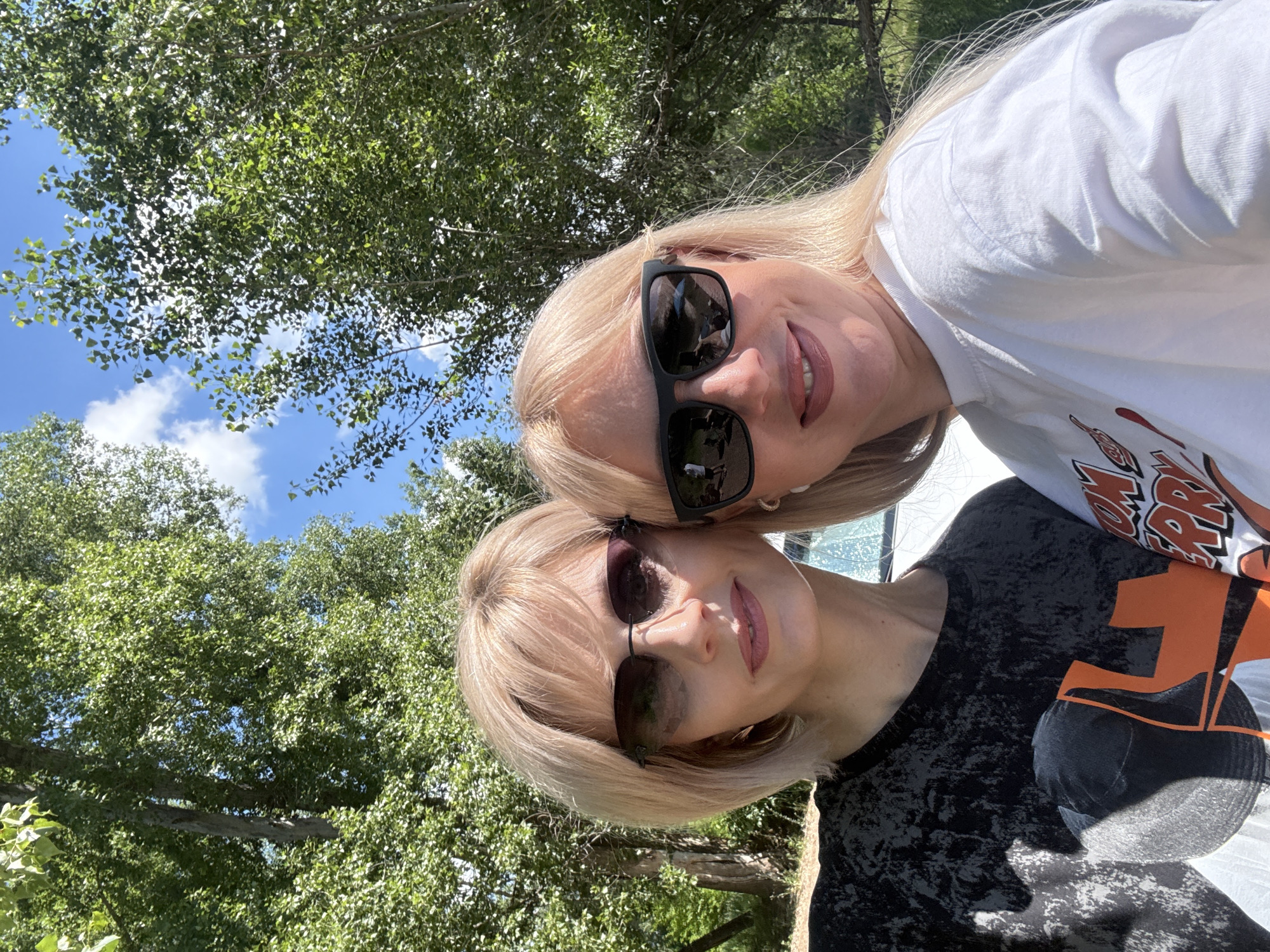 Two women, likely in their late 30s to early 40s, pose for a selfie outdoors, possibly in a park or wooded area, under a blue sky with scattered clouds. The woman on the left has short, blonde bobbed hair, wears dark-rimmed sunglasses, and a dark t-shirt with a distressed pattern and an orange graphic. She is smiling slightly with her lips parted. The woman on the right has long, blonde hair, wears dark square-framed sunglasses, and a white t-shirt with a graphic design. She is smiling broadly, with dimples visible. Both women appear to be wearing makeup. Behind them, lush green trees form a canopy, with sunlight filtering through the leaves. A portion of a white vehicle is visible between the two women, and what appears to be a fence or railing is in the background on the right. The scene suggests a casual, sunny day outing. The location is specified as Ishimbay, Russia.