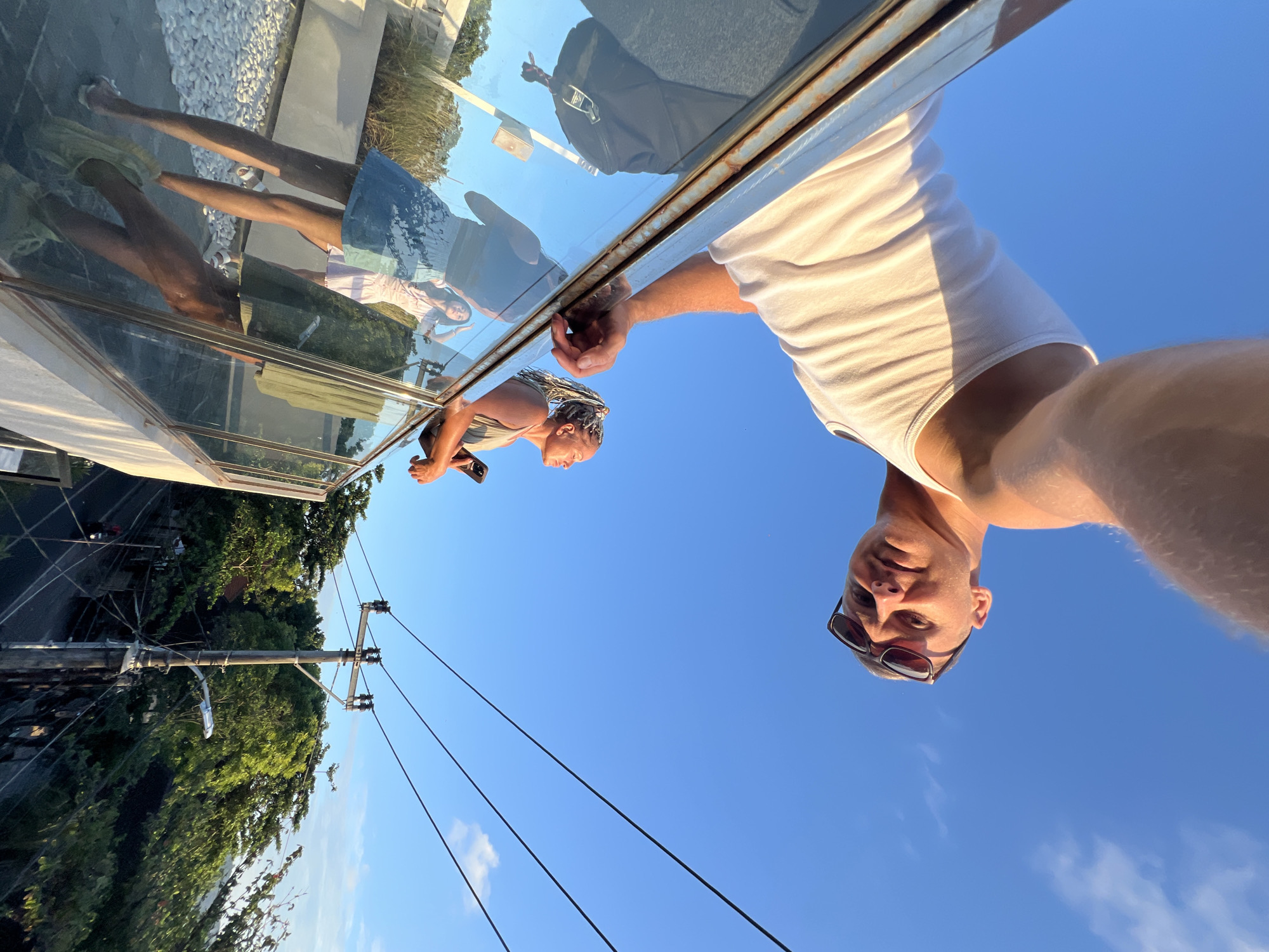 A man in a white tank top and sunglasses leans over the edge of a glass walkway, taking a selfie. Reflected in the glass are the legs of two people and the reflection of a woman looking at her phone. Below the walkway are trees, powerlines, and a road. The clear blue sky dominates the upper portion of the image. The location is Kuta, Indonesia.