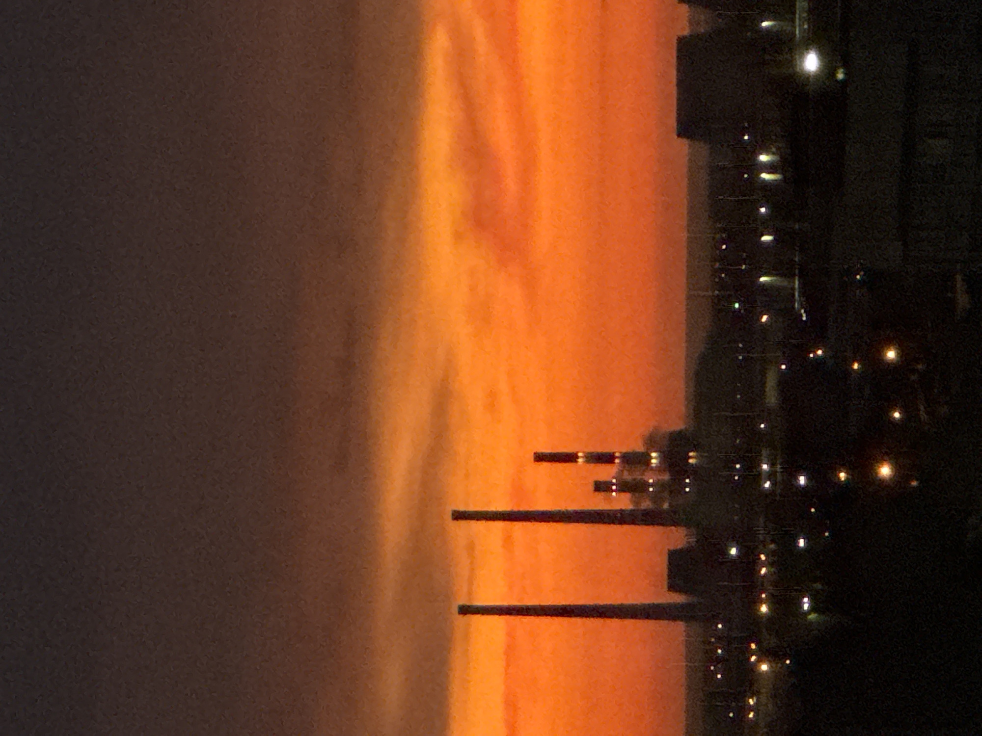 The image depicts an industrial and urban landscape at dusk or dawn, with a prominent, vivid orange and red sky dominating the middle and lower-middle sections. The upper sky is a darker gray-brown, with subtle cloud formations visible in both the dark and bright sky areas.
In the mid-ground, against the bright sky, stand several tall, dark, silhouetted industrial structures. On the left, two very tall, slender cylindrical structures, likely chimneys or smokestacks, rise prominently. To their right and slightly behind, a cluster of shorter chimneys and industrial buildings are visible, illuminated by several small, distinct yellow-white lights, suggesting operational activity.
Below the horizon line of these industrial structures, the lower portion of the image is largely dark, representing a city or industrial area at night. Numerous small, scattered points of light, appearing yellow or white, are visible throughout this dark foreground, indicating streetlights, building lights, or vehicle lights. No people are discernible. The location is an unknown city, in an unknown country.