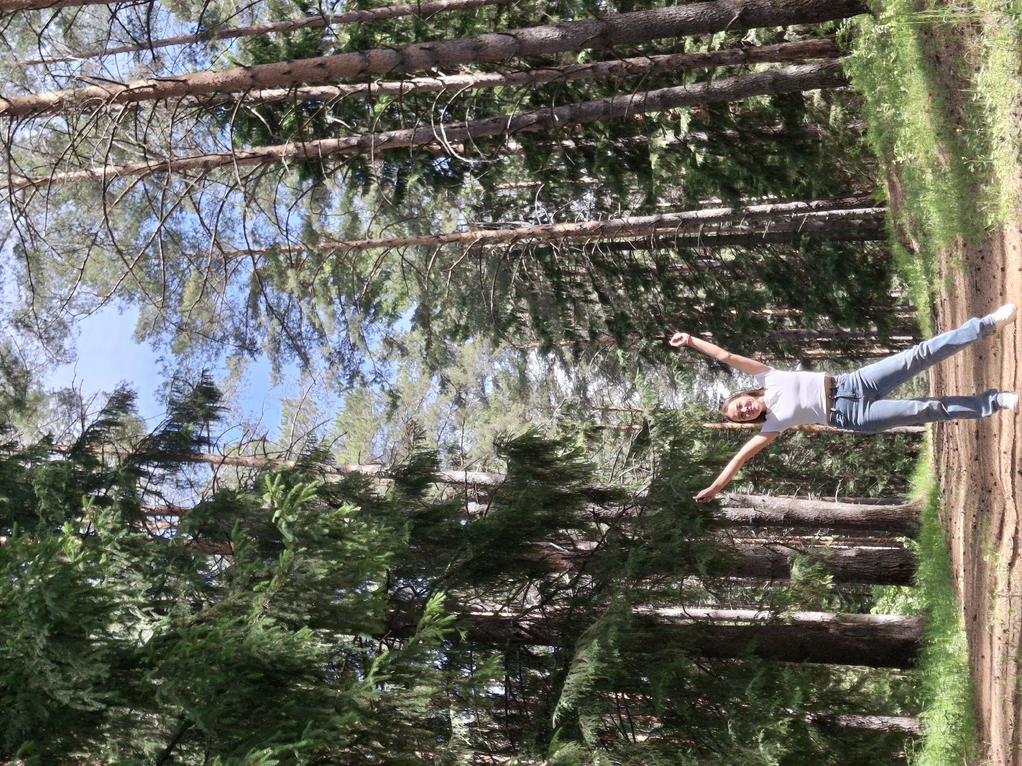 A full-length photo rotated 90 degrees depicts a fair-skinned woman standing with her arms extended outward in what appears to be a forest. The trees are tall and dense, with green foliage at various levels. The sky is visible in patches through the upper branches. The ground is covered in brown dirt and patches of green grass. The woman is wearing light blue jeans, a white t-shirt, and white shoes. The location is Прикамский, Russia.