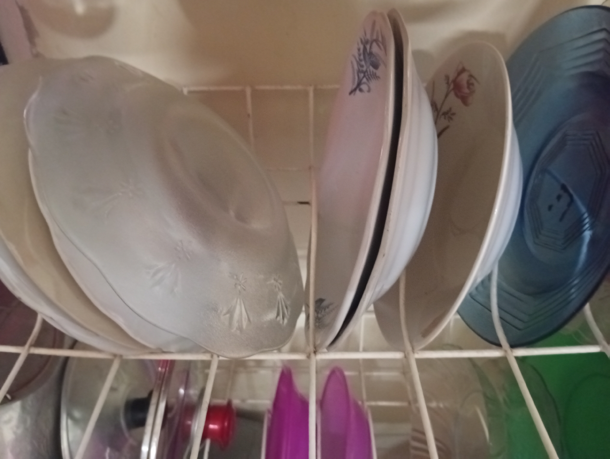 The image shows a dish rack with several plates and bowls. On the top rack, there are clear glass plates with embossed floral patterns, a pink plate with a floral design, a light-colored plate with a rose motif, and a blue plate with a geometric pattern. Below, there are various kitchen items including a pot with a red lid handle and some purple containers. The dish rack is made of white metal. The background appears to be a kitchen or dining area with a neutral-colored wall. There are no people visible in the image. The lighting suggests an indoor setting.
