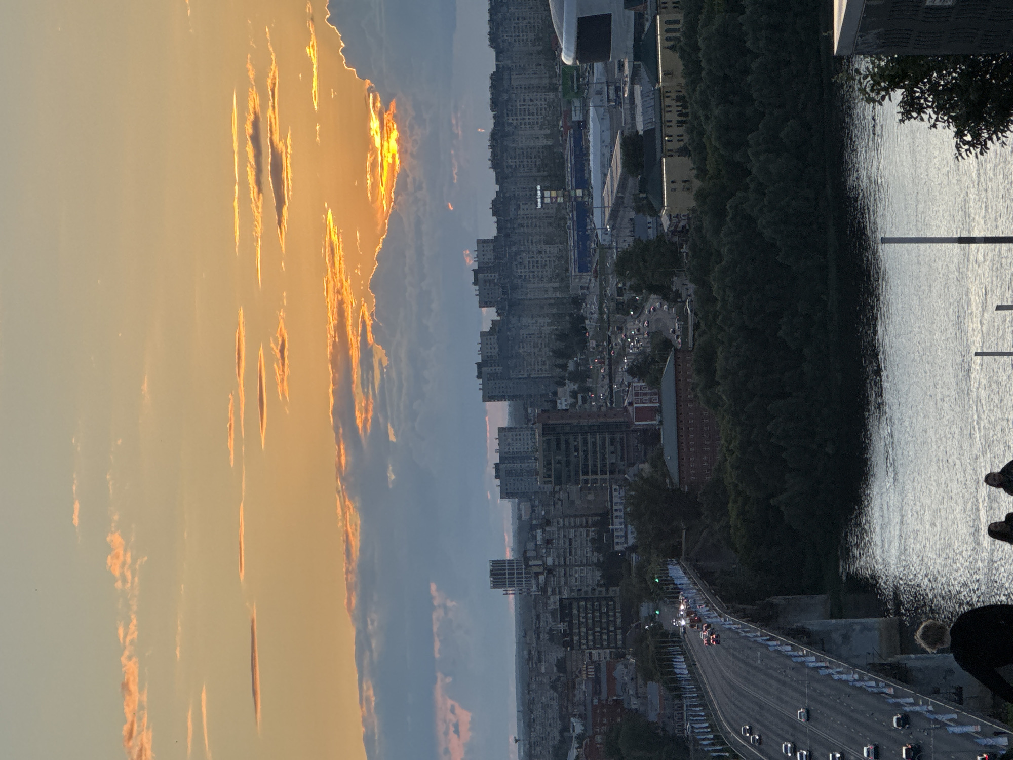 This image captures a sunset over the city of Nizhny Novgorod, Russia. The sky is filled with vibrant orange and yellow hues, with scattered clouds illuminated by the setting sun. In the foreground, a wide river reflects the sky's colors. A prominent bridge, likely the Nizhny Novgorod Cable-Stayed Bridge, spans the river, with cars driving on it. The cityscape features a mix of modern high-rise buildings and older structures, with a dense line of trees bordering the riverbank. In the lower right portion of the frame, the backs of three people are visible, looking out at the view. One person in the foreground is silhouetted against the scene.