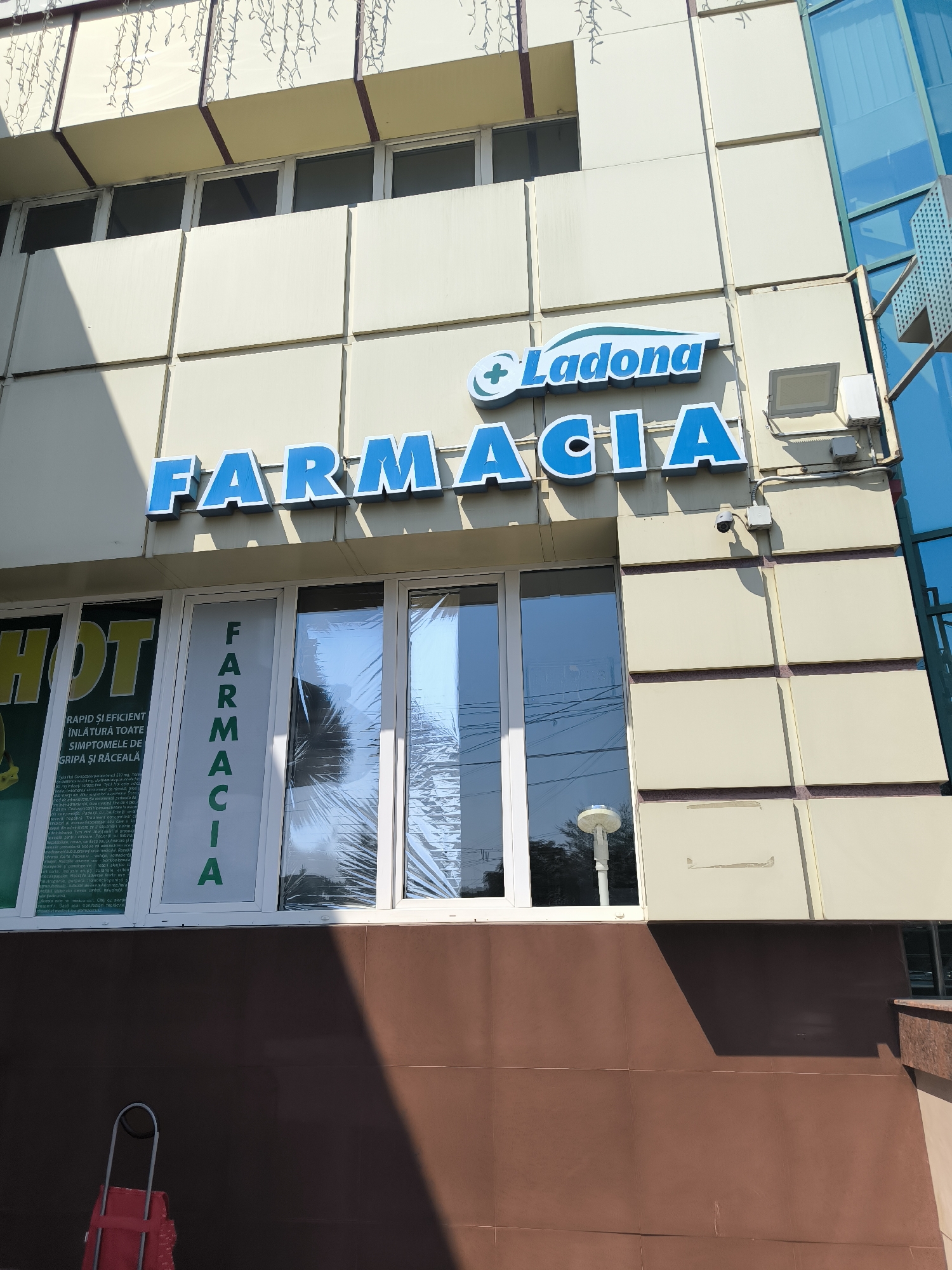 The image shows a storefront in Chișinău, Moldova. The building has a light-colored facade with several windows. Above the windows, a sign reads "FARMACIA LADONA" in blue letters. One of the windows has a vertical sign that also says "FARMACIA" in green letters. Another window has a sign with the word "HOT" in yellow letters. A red shopping cart is visible in the lower left corner of the image. A security camera is mounted on the wall above the windows. The sky is visible in the reflection of the windows.