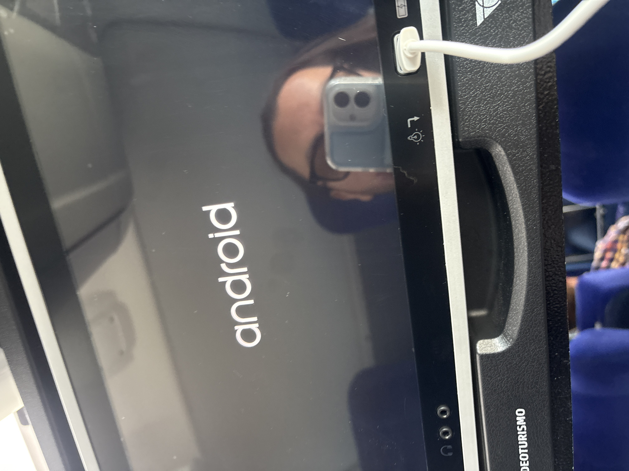 An indoor scene captured at a slight angle shows a dark digital screen, possibly an in-vehicle entertainment system, displaying the white "android" logo on a black background, indicating it is booting up. A person's reflection, including their head, shoulders, and a smartphone with a multi-lens camera (resembling an iPhone in a clear case), is prominently visible on the screen's surface. A white cable, likely a charging cable, is plugged into the top right edge of the device, extending upwards and to the right.
The screen is framed by a black, textured border. On this border, near the top right, a small lightbulb or lamp icon is visible, suggesting a control for illumination or a reading light. A power or battery icon is also partially visible above the charging cable. Further down the right side of the frame, the word "VIDEOTURISMO" is printed vertically in white text, implying the device is part of a video tourism system. Below the screen, three small circular indents or ports are partially visible on the frame.
In the background, out of focus, are parts of dark blue upholstered seats, reinforcing the setting as inside a passenger vehicle, likely a bus or train. The image does not provide cues for the time of day or specific weather conditions. The presence of "VIDEOTURISMO" and the seating arrangement align with the provided location context of Valle de Chalco Solidaridad, Mexico, suggesting this is a public transport or tour vehicle within the region. The activity is primarily the screen booting up and the user capturing its reflection.