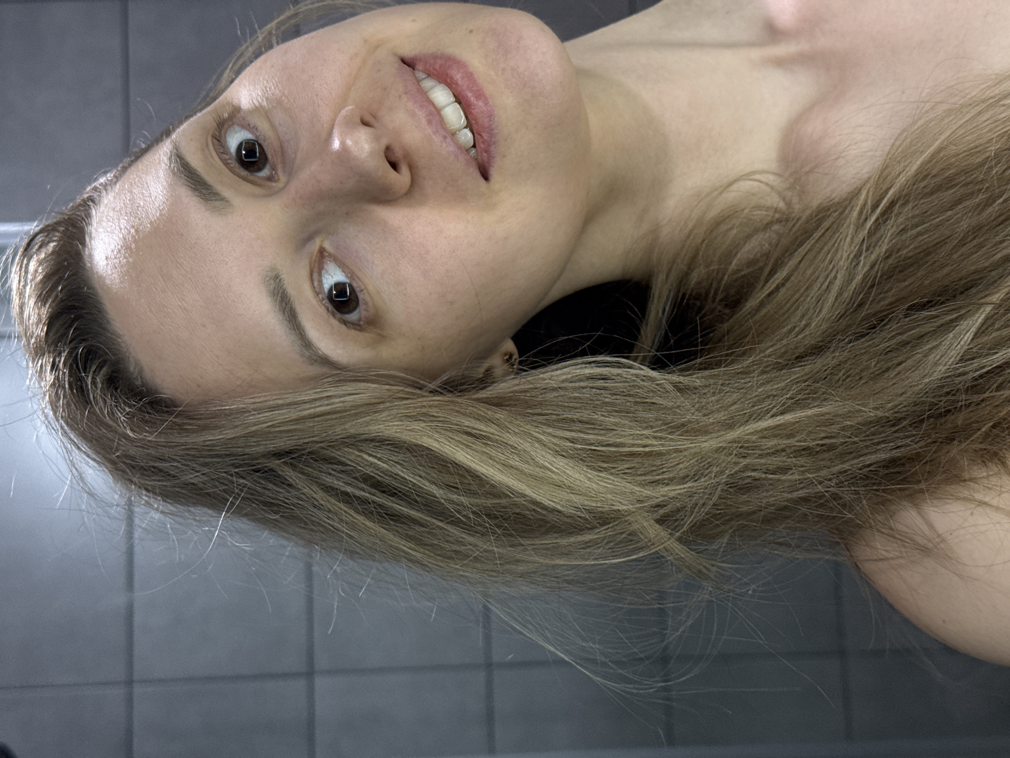 A woman with light brown hair and brown eyes is lying down. She is fair-skinned and has a slight smile. The background is a gray tiled surface. The image does not contain enough information to determine a location in the city of Chaikovsky, Russia.