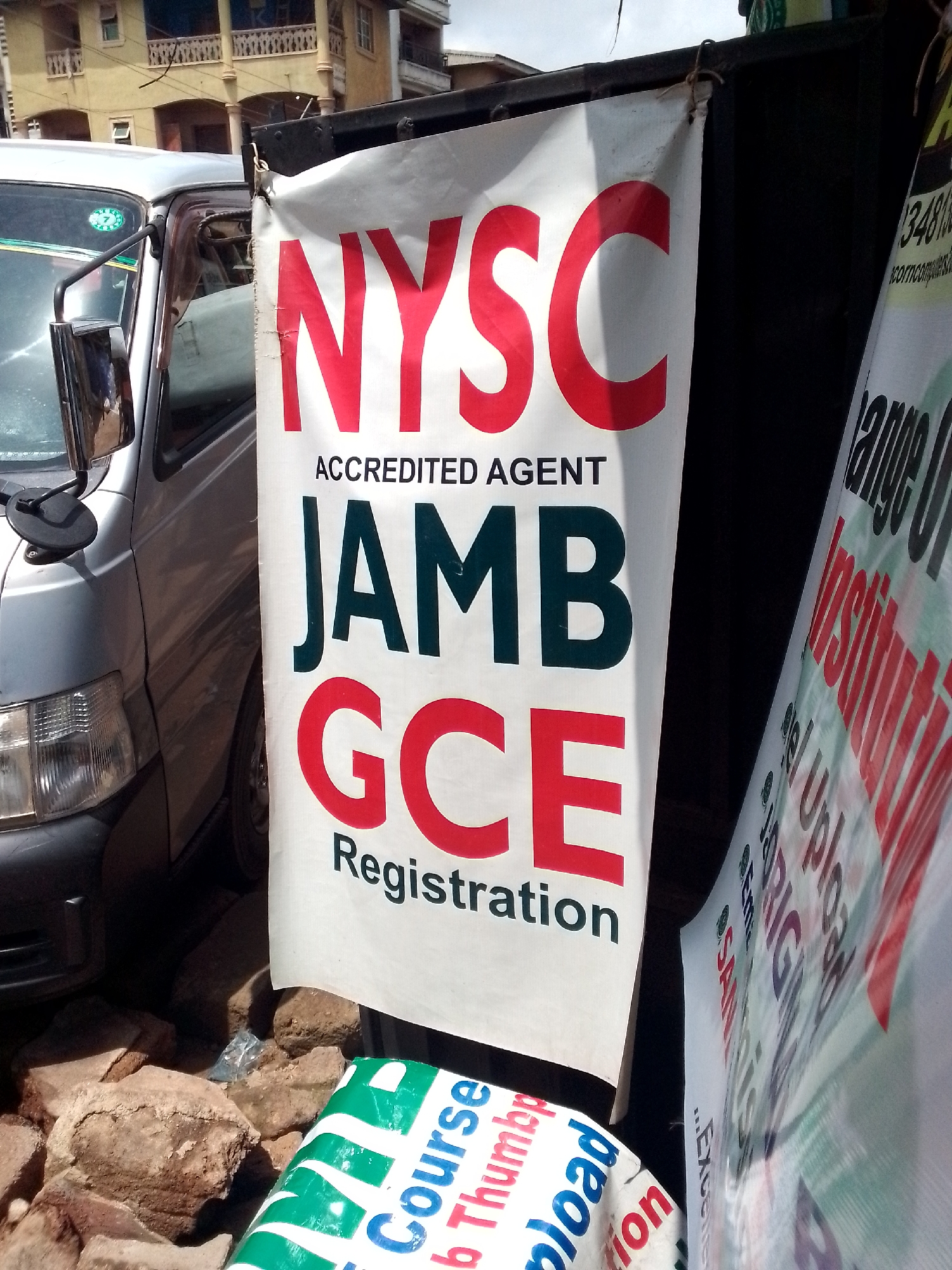 A close-up, eye-level, outdoor shot captures a white banner displaying red lettering for "NYSC", "JAMB", and "GCE", with smaller black text reading "ACCREDITED AGENT" and "Registration". The banner is hung from a dark, possibly metal, surface, with a silver van partially visible to the left. Rocks are piled on the ground below the banner. To the right, another banner with red and green lettering is visible, its content partially obscured. A building with balconies is in the background. This scene is located in Nkpor, Nigeria.