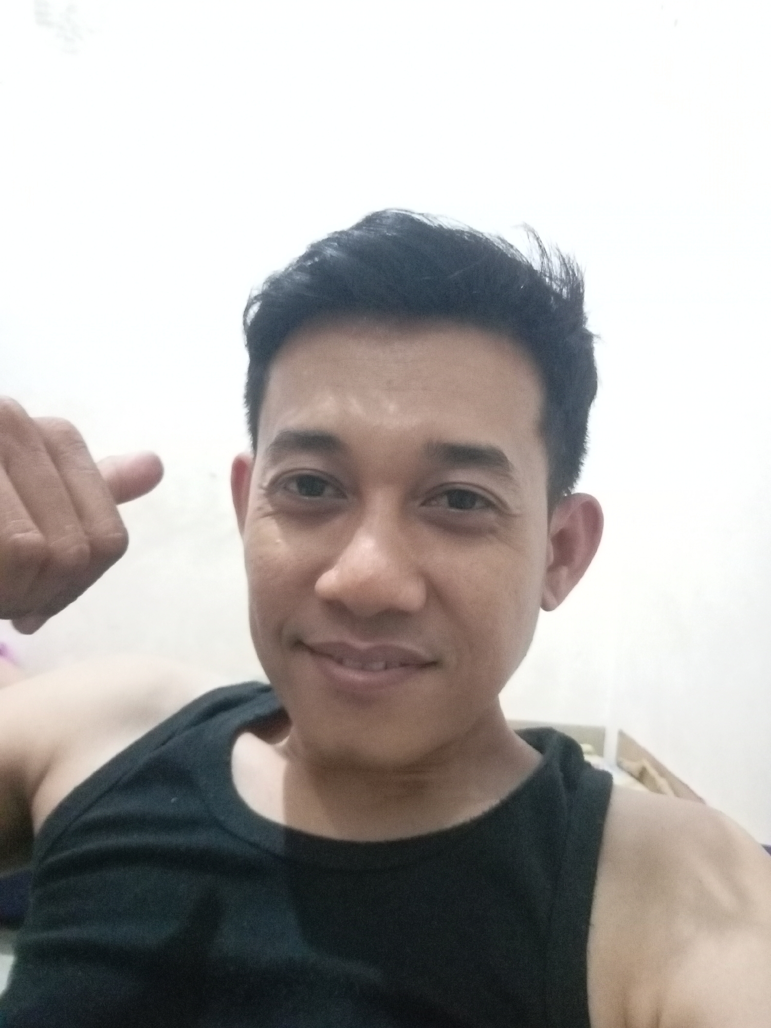 A man with dark hair and dark eyes, wearing a black tank top, is looking at the camera and pointing his left thumb up. He has a slight smile on his face. The background is a plain white wall. The scene appears to be indoors, possibly in a home in Batam, Indonesia, though no specific location details are visible in the image.