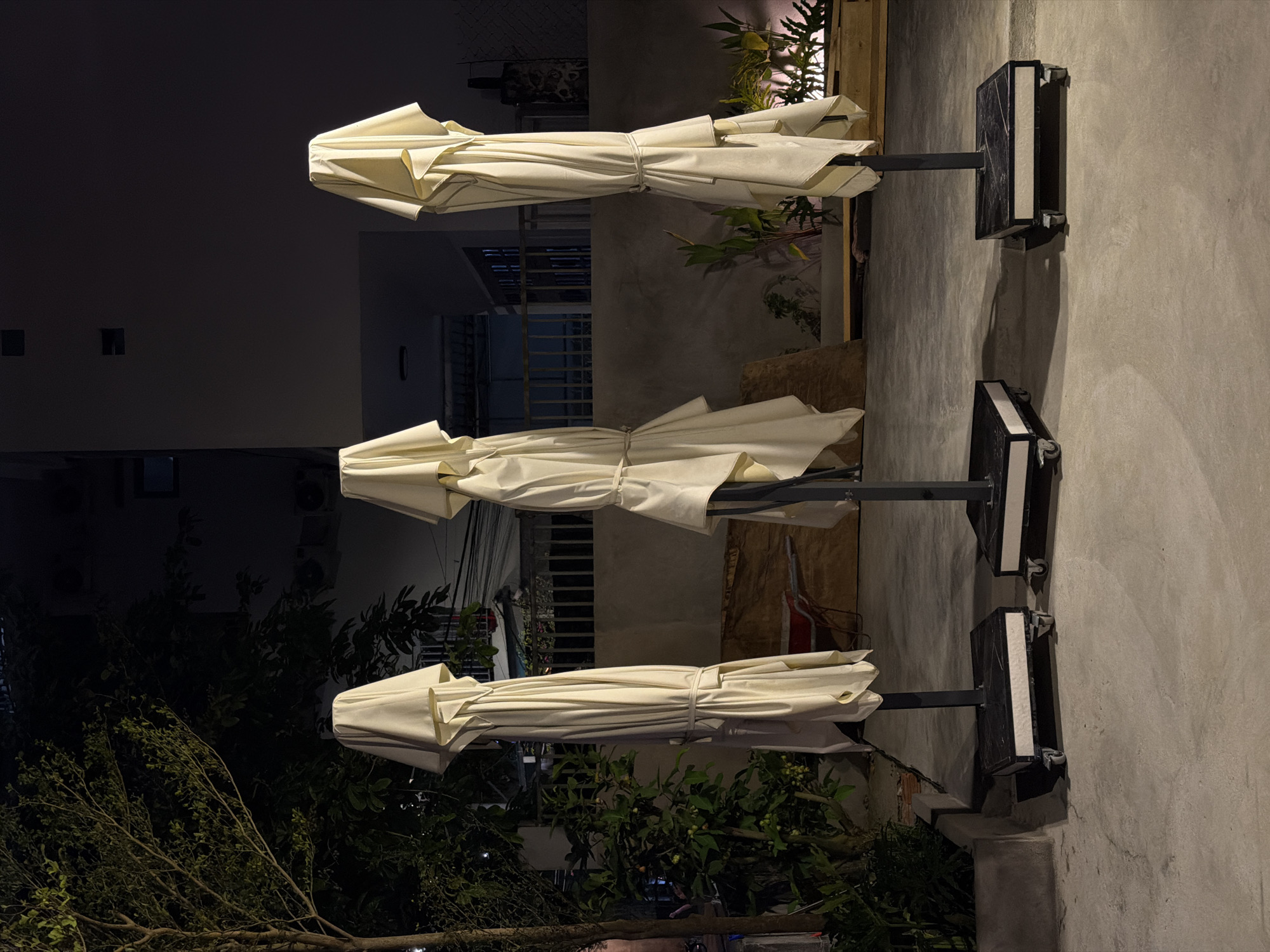 Three folded white umbrellas are positioned side-by-side outdoors, each secured to a black metal stand. The stands are affixed to a light gray concrete wall. Behind the umbrellas are various plants and a building with windows. The lighting suggests it is nighttime. The location is likely Đà Nẵng, Vietnam, given the architectural style and flora.
