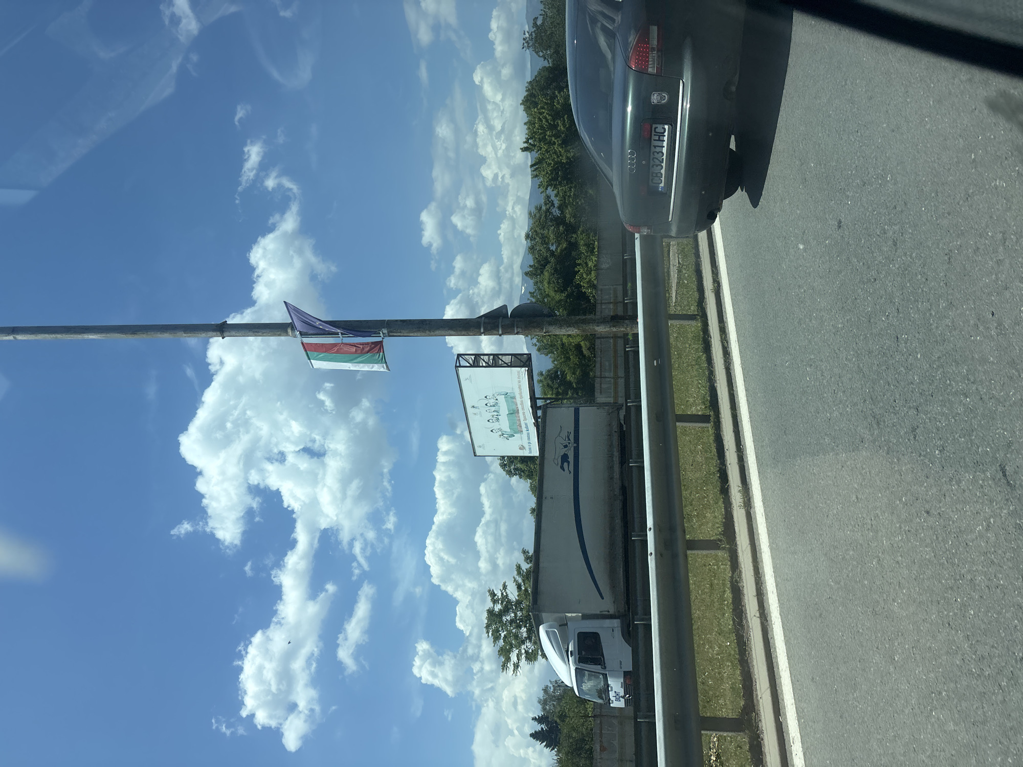 The image captures a roadside scene under a bright blue sky with scattered white clouds. In the foreground, a dark-colored Audi sedan is visible, displaying the license plate "CB 3231 HC". Adjacent to it is a guardrail and a portion of a highway. Further in the background, a white truck with a blue stripe and two running dogs logo is driving on the highway. A billboard is also present, depicting a group of people, and a Bulgarian flag is hanging from a lamppost. The surrounding environment includes trees and mountains in the distance. The location is identified as the city of German, Bulgaria.
