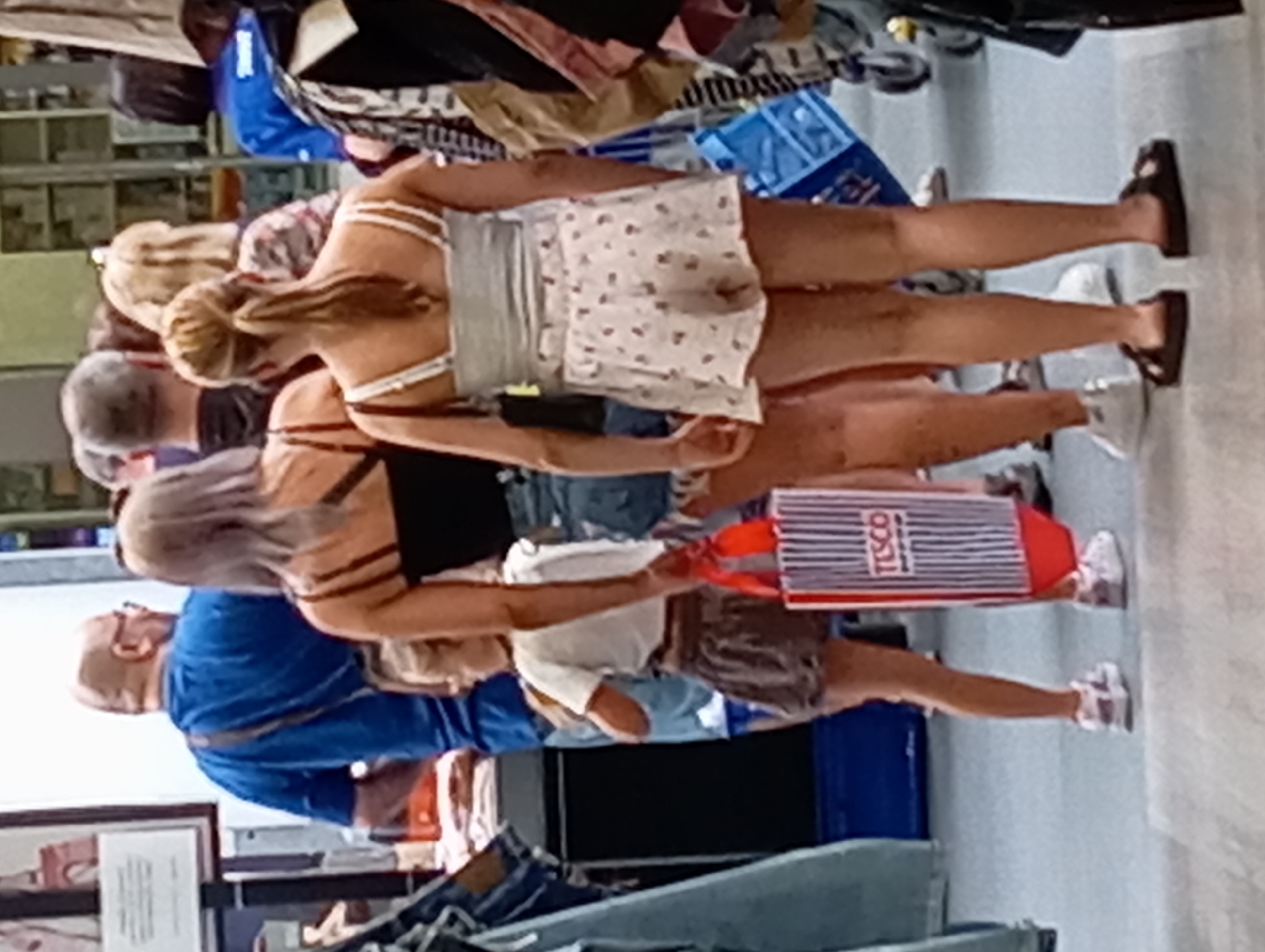 The image depicts a scene from a commercial environment, likely a supermarket or a department store, given the presence of shopping bags and displays of goods. The floor appears to be tiled. In the foreground, two women and a young girl are visible, all facing away from the camera. The woman on the left wears a black top and denim shorts, while the woman on the right is in a light-colored top and a printed skirt. The young girl, positioned between them, wears a white t-shirt and a dark skirt. A Tesco branded shopping bag is held by the woman on the left, with the "TESCO" logo clearly visible. To the left of this group, a portion of a person wearing a blue t-shirt and carrying a bag over their shoulder is visible. In the background, other shoppers and store shelves are blurred. The lighting suggests an indoor environment. The context of Nitra, Slovakia, is not directly evident from visual cues in the image itself.