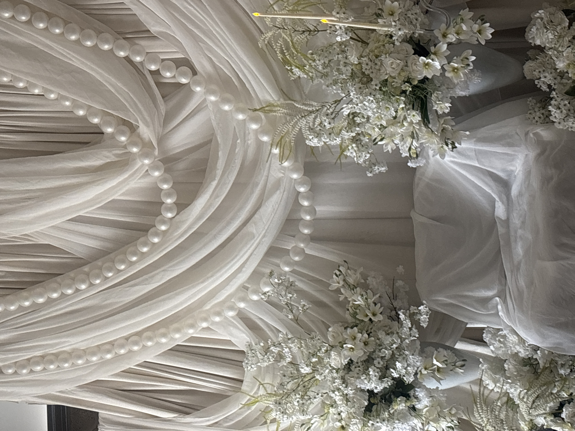 The image shows a wedding decoration, likely indoors. White drapery is arranged in flowing patterns, adorned with strands of pearls. White floral arrangements with greenery accent the fabric. The background is out of focus, but the setting appears to be a room decorated for a special event. The image has no indications of its exact location within Yerevan, Armenia. No people are visible.