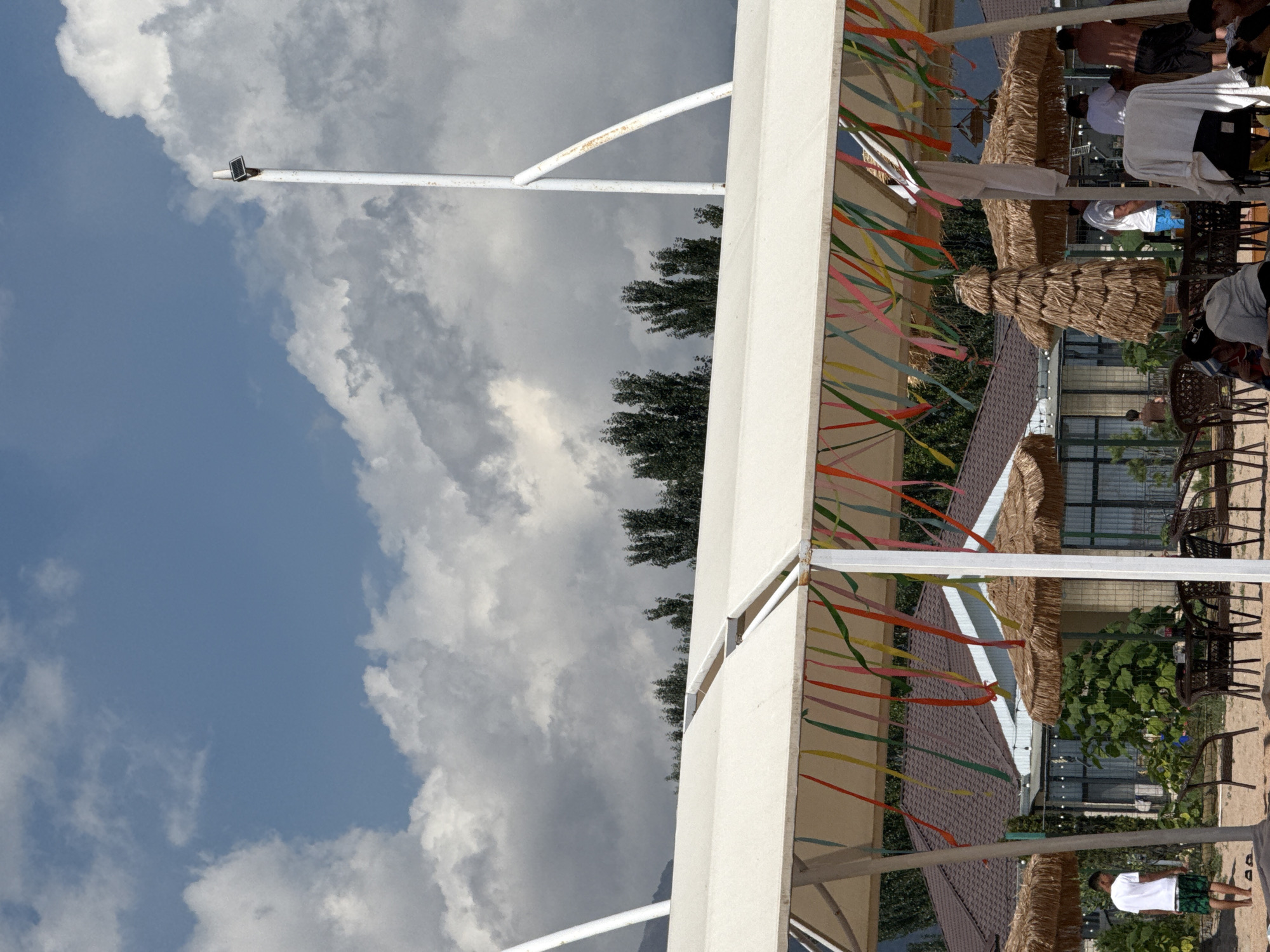 In Sary-Oy, Kyrgyzstan, a beachside area is depicted under a cloudy sky. In the foreground, a white awning with colorful streamers provides shade over a seating area on a sandy ground. Several straw umbrellas are visible, as are tables and chairs. A young man in a white t-shirt and green shorts stands on the sand, facing away from the camera. In the background, a building with a brown tiled roof is partially obscured by lush green foliage. Further back, trees stand against the backdrop of a mountain range. A white pole with a solar panel is prominently positioned on the right side of the frame, reaching towards the sky. People are scattered throughout the scene, some appearing to be near the building, while others are under the umbrellas.