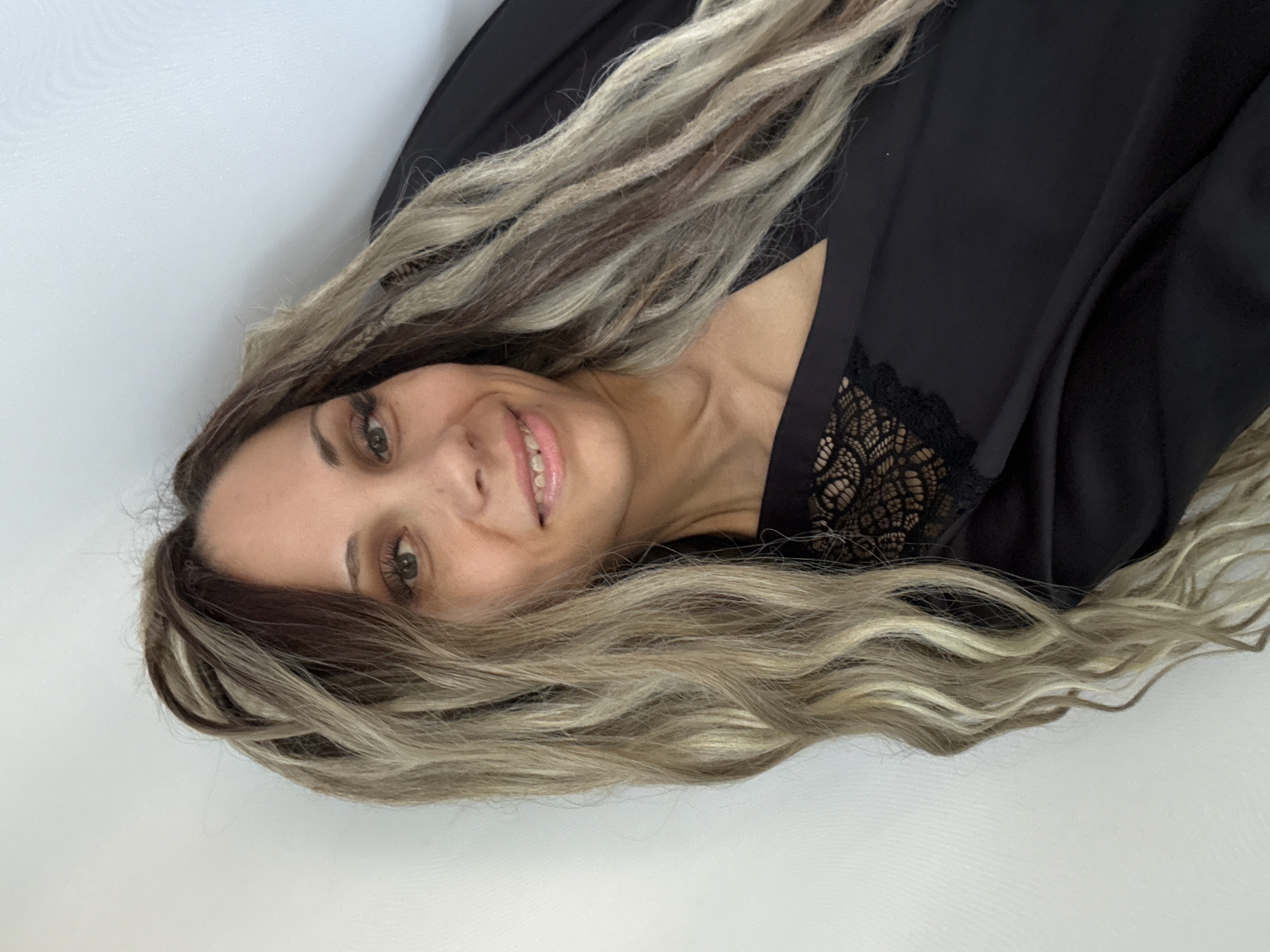 A woman with long, blonde and brown wavy hair smiles at the camera. She is wearing a black robe with lace accents on the neckline. The background is a plain white wall. The image appears to be a selfie, taken indoors. There is no information to suggest this image is located in Rusanovo, Russia.