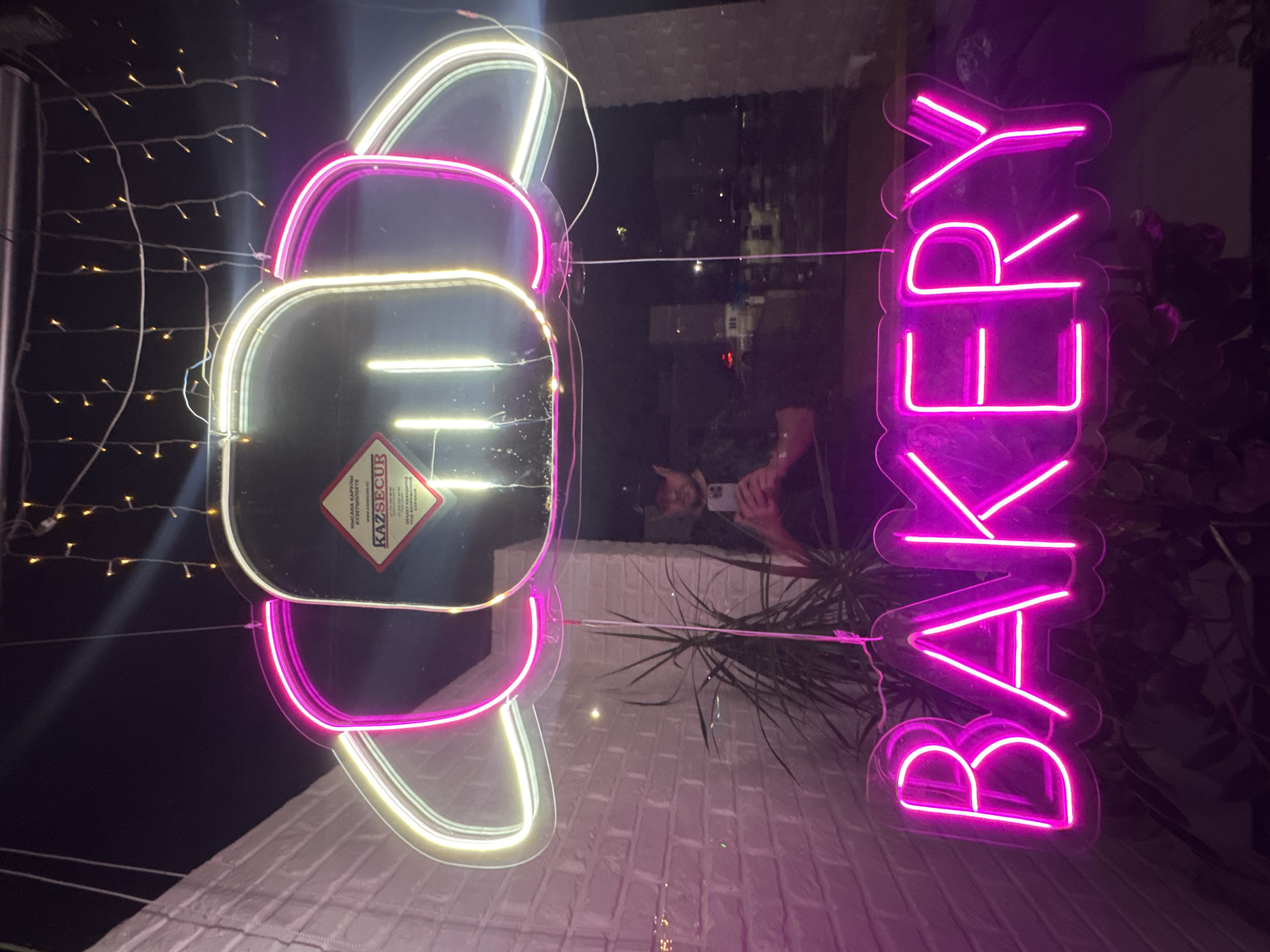 An exterior shot taken at night of a bakery in Almaty, Kazakhstan, is captured. A neon sign, shaped like a croissant and lit with pink and white lights, hangs in the window, reflecting the interior. Below the croissant sign, a bright pink neon sign spells out "BAKERY." A man wearing a dark baseball cap is visible in the reflection, holding a smartphone. The background shows a brick wall on the left, and a blurry cityscape through the window on the right. A potted plant with long, thin leaves is positioned in front of the signs.