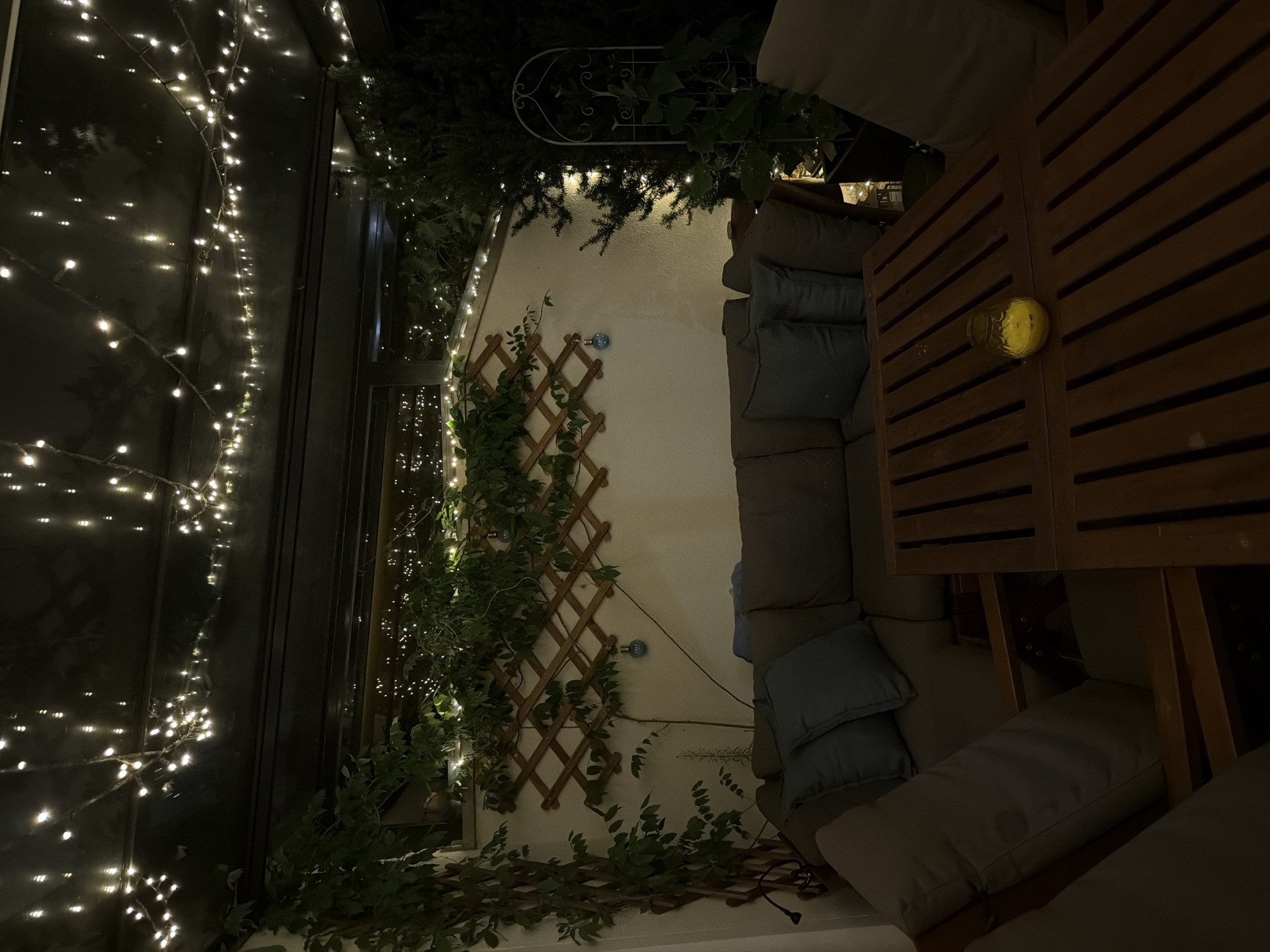 The image depicts a dimly lit outdoor patio area at night, likely in Poing, Germany. The space is adorned with numerous strings of warm white fairy lights draped across the ceiling, creating a festive and cozy atmosphere. In the foreground, a wooden slatted coffee table holds a single yellow candle in a glass holder, its light casting a soft glow. A sectional sofa with dark gray cushions and lighter gray accent pillows is arranged around the table, suggesting a comfortable seating area. Behind the sofa, a white wall serves as a backdrop, decorated with a wooden lattice against which green vines are climbing. Two small blue decorative spheres hang from the lattice. To the right of the sofa, a dark green evergreen tree is visible, also illuminated by fairy lights. Further to the right, a black metal garden trellis stands, partially obscured by foliage. The overall impression is a tranquil evening setting, possibly a balcony or enclosed patio, prepared for relaxation or socializing.