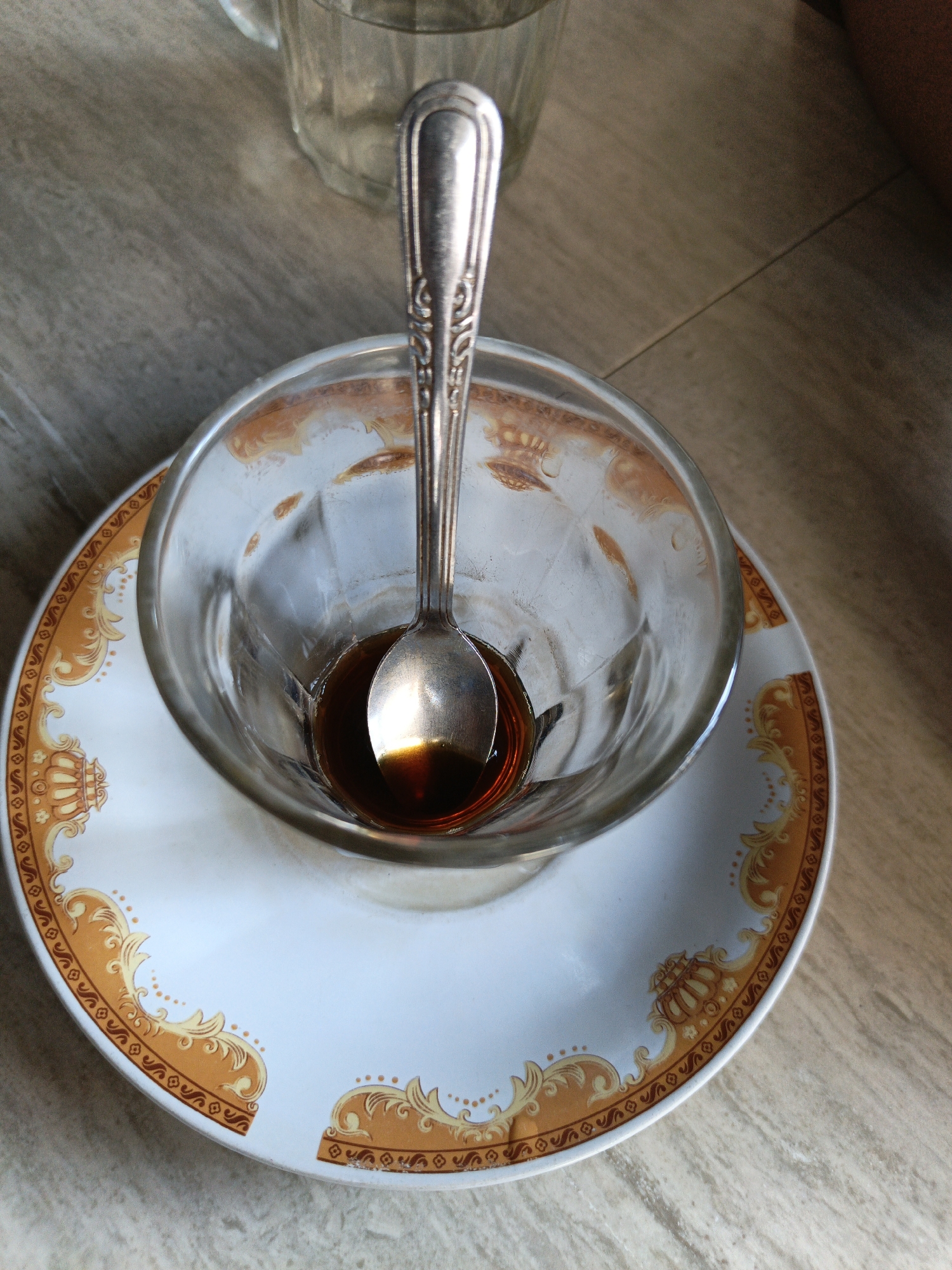 A used glass cup sits on a white saucer with a gold decorative pattern. Inside the cup is a silver spoon resting in a small amount of dark liquid. The scene is on a light-colored wooden surface. The location is unspecified, although the user states it's in Lhokseumawe, Indonesia. Other items on the table include a glass and the arm of a person. The scene depicts the remnants of a beverage.