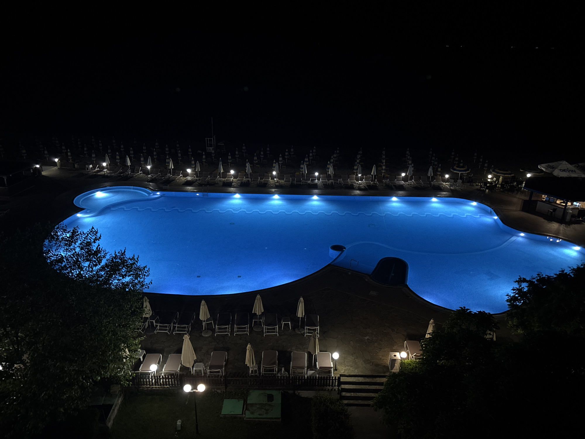 The image is an aerial view of an outdoor swimming pool at night, likely in Varna, Bulgaria. The pool is large and illuminated with vibrant blue underwater lights. Surrounding the pool are rows of empty lounge chairs and closed umbrellas, suggesting the area is prepared for daytime use. In the background, a beach with more umbrellas and chairs can be seen, along with a small building that appears to be a bar or restaurant with outdoor seating. Streetlights and spotlights illuminate the area around the pool and beach. Trees partially obscure the view on the left and right sides of the image. The overall scene is tranquil and empty, with the glowing pool being the central focus.