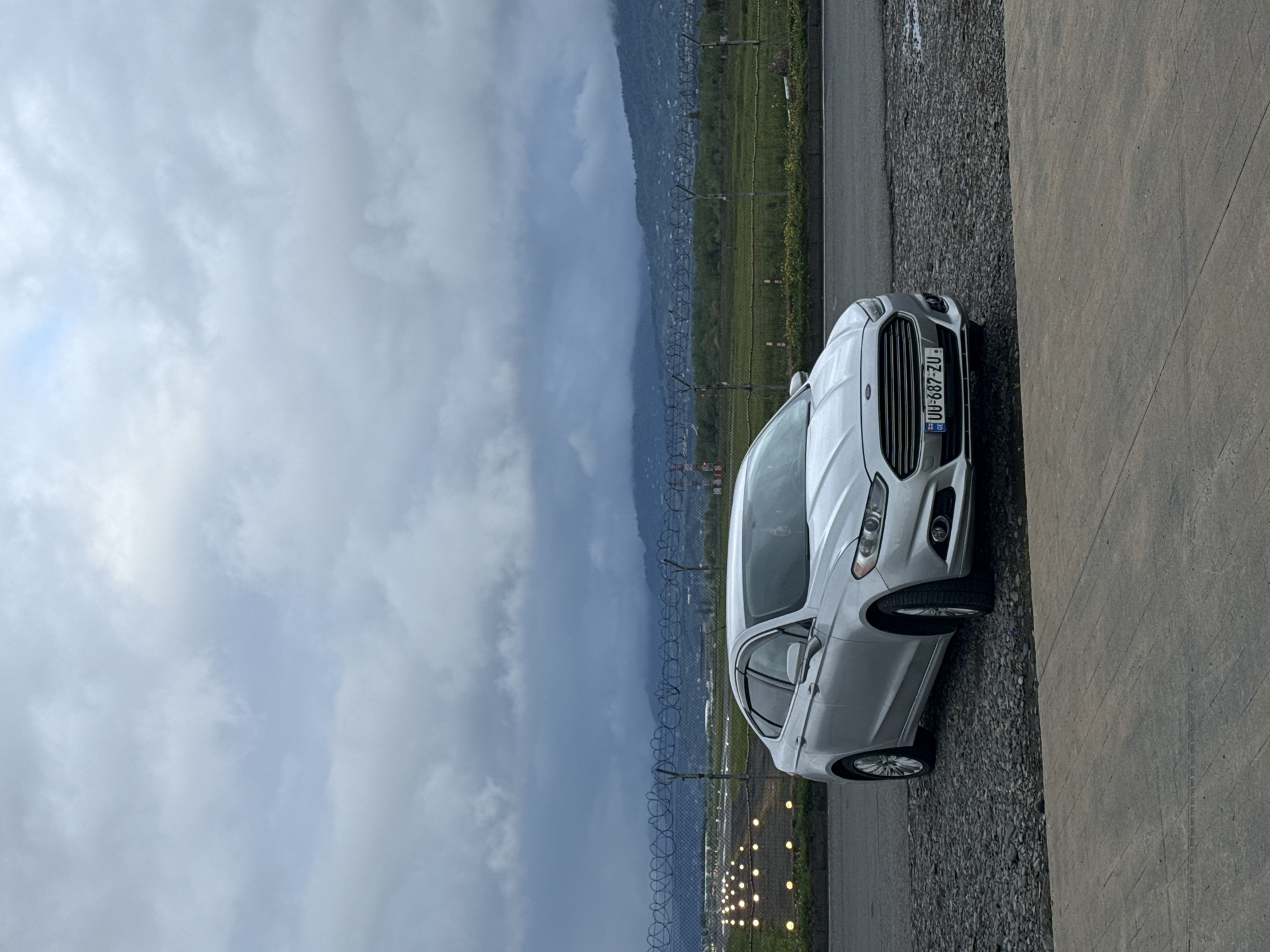The image shows a silver Ford Fusion with license plate "UU-687-ZU" parked near the Batumi International Airport in Georgia. The car is adjacent to a concrete surface and a gravel area. In the background, there's a mountain range obscured by clouds, as well as a fence bordering the airport runway, which is marked by runway lights. The sky is overcast, suggesting cloudy weather.