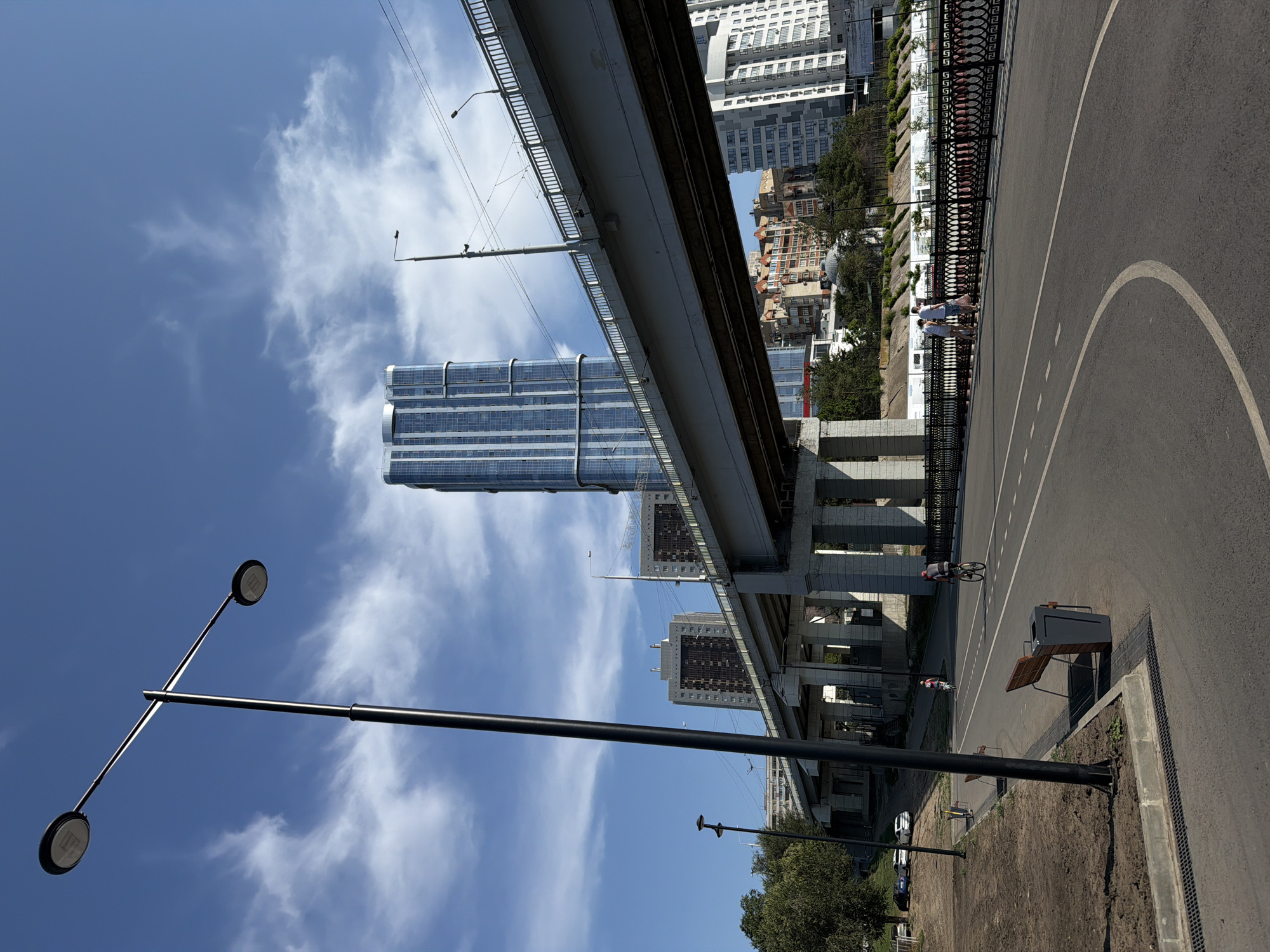 A wide-angle, eye-level shot captures a sunny day in Saratov, Russia, featuring a pedestrian and cyclist path alongside a river with a modern bridge and urban skyline. In the foreground, a paved path curves to the right, marked with white dashed lines, and is bordered by a black metal fence on the right and a grassy area with a dark grey trash receptacle and a wooden bench on the left. A tall, black lamppost with two lights stands to the left of the path.
Further back, two individuals are seen walking near the fence, while two cyclists are on the path, one further ahead than the other. The bridge, a prominent structure, spans across the scene from the left middle ground to the right middle ground, supported by concrete pillars. Beyond the bridge, a cluster of buildings forms the city's skyline, including a tall, modern skyscraper with a blue glass facade. The sky above is bright blue with scattered white clouds. The environment suggests a developed urban area with recreational spaces.