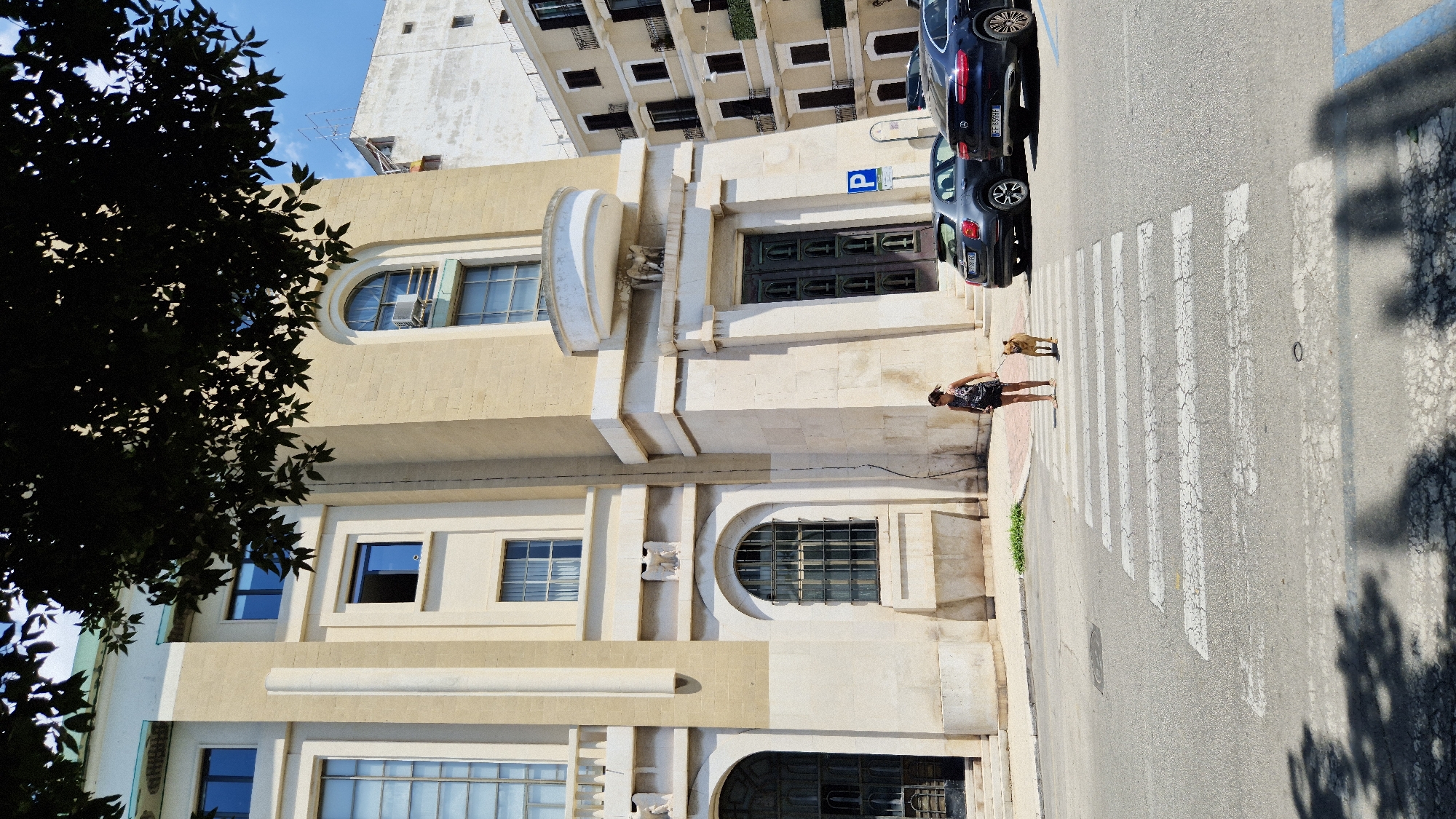 In Taranto, Italy, on a bright, sunny day, a woman walks a light brown dog across a pedestrian crossing in front of a large, light-colored building with classical architectural elements. The woman is wearing a dark, patterned sleeveless top and dark shorts, and the dog is on a leash. Two cars are parked on the right side of the street, one dark blue sedan and one gray hatchback. A blue and white parking sign is visible behind the cars. The building features large windows, columns, and decorative elements. The sky is clear blue with scattered white clouds, and a tree in the foreground casts a shadow on the street.