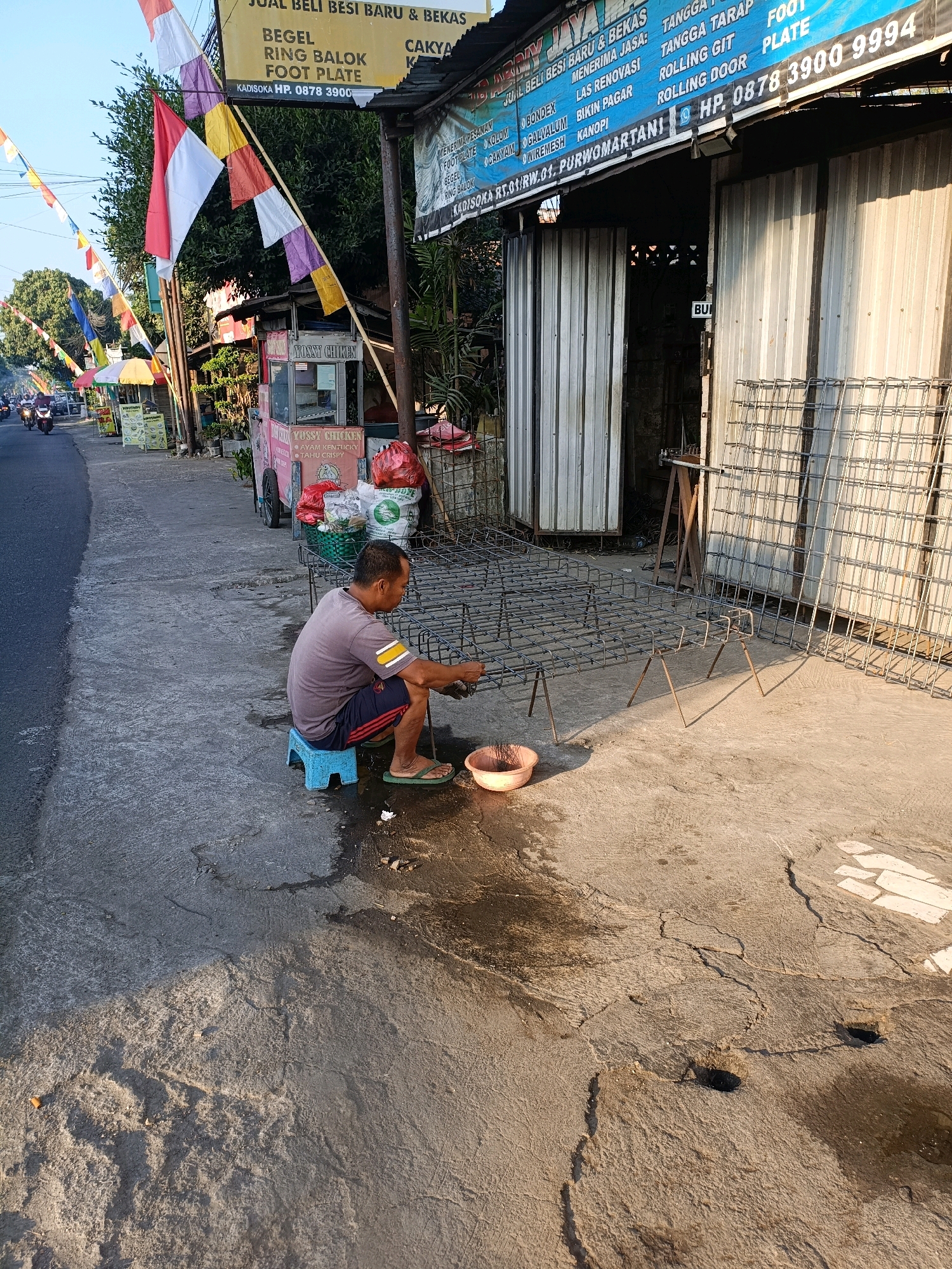 A man is sitting on a blue plastic stool on a concrete sidewalk in Maguwoharjo, Indonesia. He is wearing a grey t-shirt and dark shorts with red stripes. The man is working on a structure made of metal rebar, which is laid out on the ground and supported by metal legs. In front of him is a small orange bowl with some liquid and a brush. A large metal rebar structure is leaning against a building to his right. The building has a corrugated metal facade.
To the left of the man, there is a sidewalk with several food carts. One cart has "YUSSY CHICKEN" written on it in pink lettering. Behind the food carts, there are trees and colorful flags strung across poles. In the background, a street with vehicles is visible. The lighting suggests it is either early morning or late afternoon. The ground around the man is wet and stained.