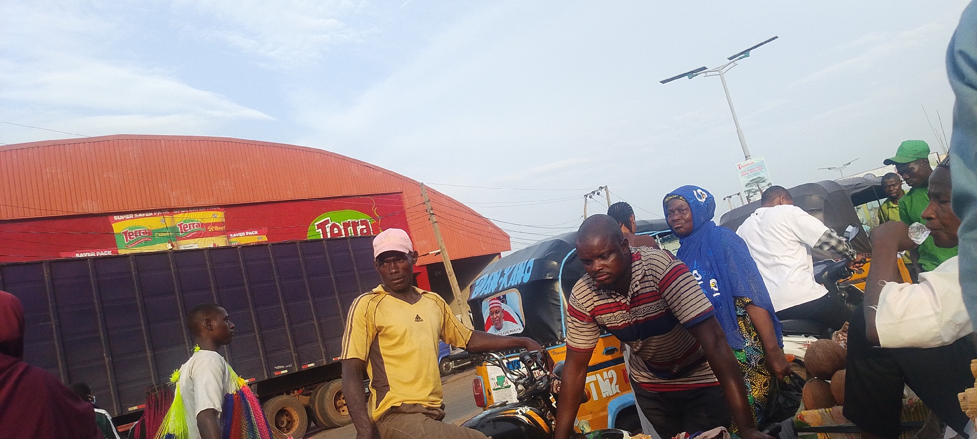 This image captures a bustling street scene in Lokoja, Nigeria, under a bright, overcast sky, suggesting daytime. The setting appears to be a market or commercial area.
In the background, a large red building with an orange corrugated iron roof dominates the left side. Adorning the red wall is a large billboard advertising "Terra" brand products, likely food items, with "SUPER SAVER PACK" visible multiple times in yellow and green lettering, and the word "Beef" also partially discernable. In front of this building, a dark blue cargo truck is parked or moving slowly.
The foreground and midground are filled with people and vehicles. A man wearing a yellow Adidas t-shirt and a pink baseball cap is seated, possibly on a motorbike or the front of a tricycle, looking towards the viewer. To his right, another man in a striped shirt is leaning over a motorbike or tricycle. A woman in a blue hooded head covering (hijab) is visible behind them, looking to the right. Further right, a man in a green cap and green shirt is seen, along with other individuals. In the lower right, a small pile of coconuts or similar round objects rests on a stand.
A prominent yellow and blue tricycle (locally known as Keke Napep) is central to the right side of the image, with "DAN KANG" written on its side. A picture of a man, possibly a local figure or politician, is displayed on the tricycle's front. Behind the vehicles, a street light pole with solar panels and a sign advertising "Dentist" and "Daily" services are visible. In the far left foreground, a person holds colorful, striped fabric or plastic items, possibly merchandise. The overall impression is one of everyday commerce and transit in a vibrant urban environment.