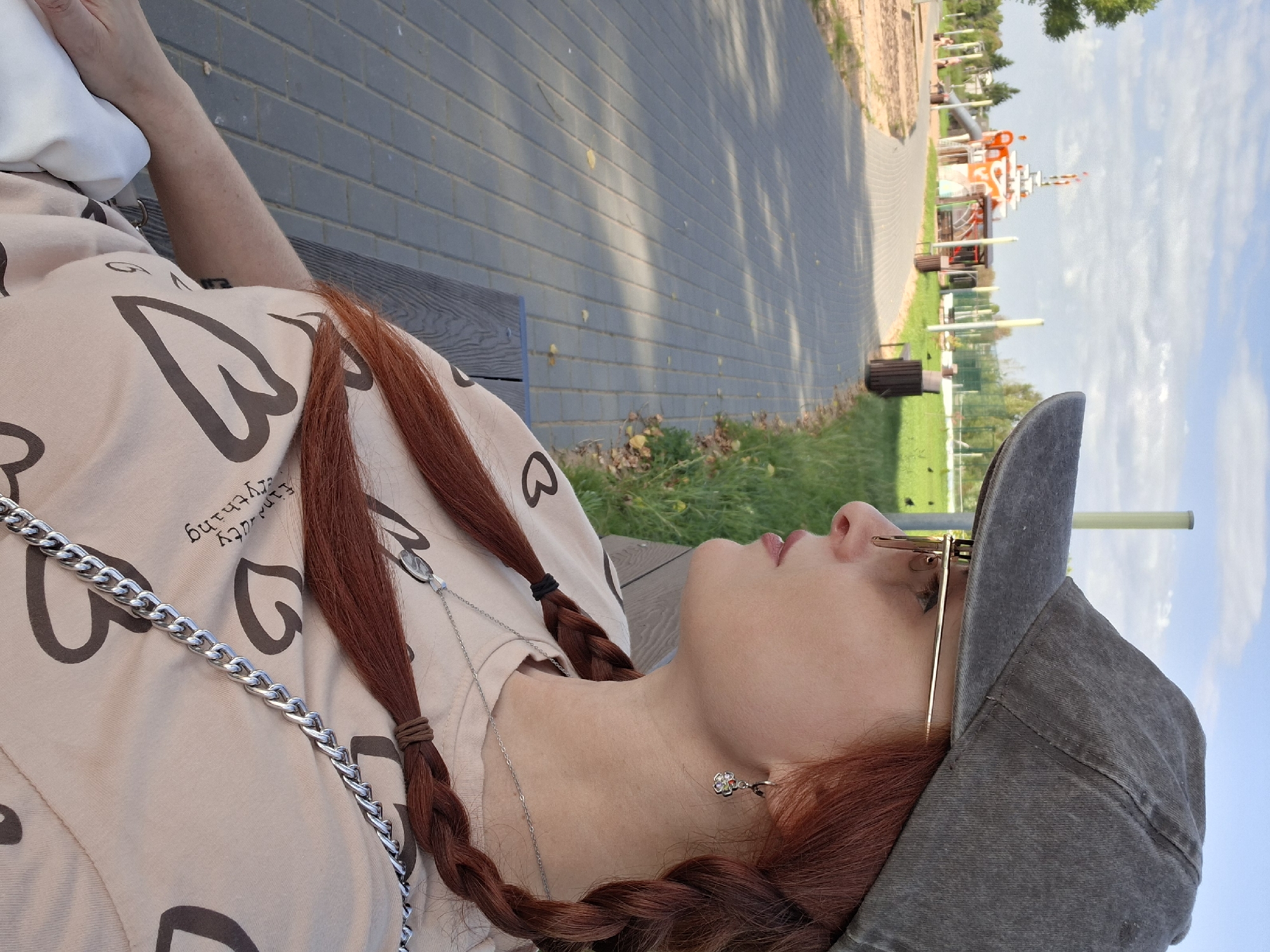 A young woman with auburn hair in braids, wearing a grey baseball cap and a light-colored t-shirt with heart patterns, sits on a bench outdoors. She is looking to her right. The background shows a park with a paved walkway, grass, trees, and a playground structure. The location is identified as the city of Myadzyel, Belarus.
