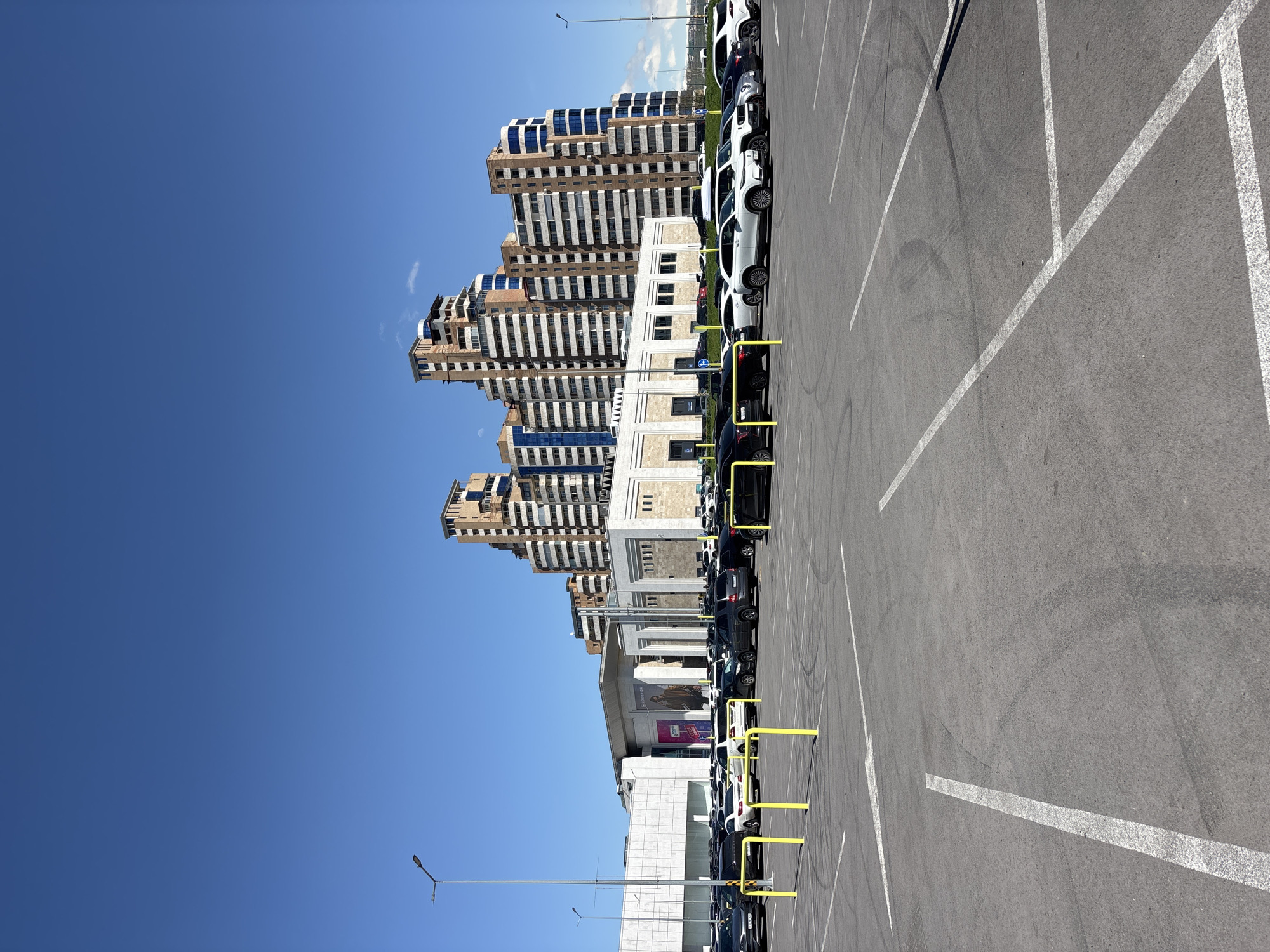 The image shows a parking lot in Yerevan, Armenia, adjacent to a building complex. The parking lot is paved with gray asphalt and marked with white lines, delineating parking spaces. Cars, primarily black and white SUVs, are parked in a row along the building side of the lot. Yellow barriers or bollards are positioned along the parking spaces. In the background, there are multi-story buildings featuring a layered architectural design, with tan-colored facades and dark windows. The sky above is clear and blue. The photo is taken during daylight.