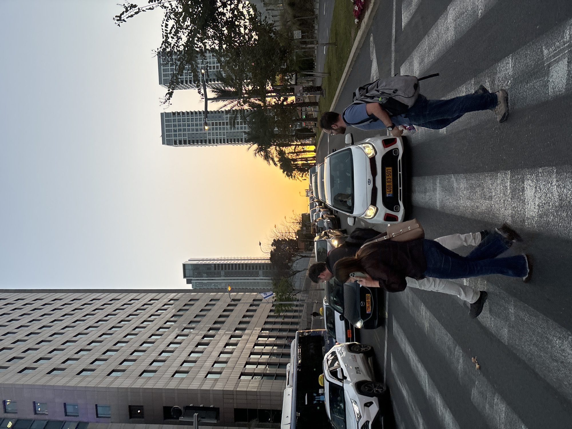 The image depicts a street scene in Tel Aviv, Israel, at what appears to be either sunset or sunrise. The sky is a gradient of yellow and light blue. Several tall buildings are visible in the background. A line of vehicles, including cars and taxis, are stopped in traffic on the road. Pedestrians are crossing the street at a crosswalk. One individual is wearing a blue shirt and jeans with a grey backpack. Another is wearing a dark shirt and jeans and appears to be holding a phone. The license plates on the cars are typical Israeli license plates.