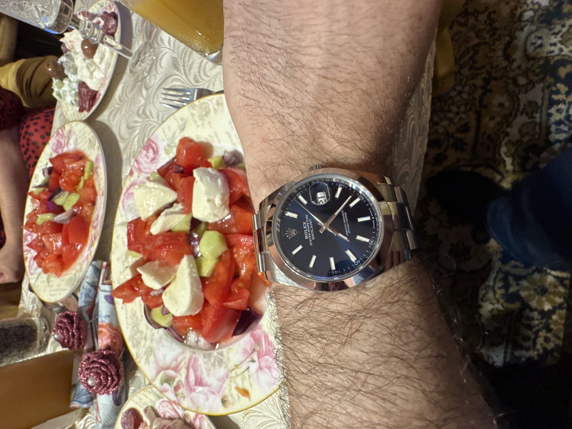 The image shows a close-up view of a hairy arm wearing a Rolex Datejust watch with a blue dial. The watch face displays the date as the 18th. Beside the arm are plates of salad with tomatoes, cucumbers, and white cheese on a tablecloth with floral patterns. Other dishes are visible in the background, along with a glass of orange juice. Part of a person's leg with a red patterned garment is visible on the left. Part of a shoe can be seen on the right. The image's context suggests a meal setting, likely at home. It is not possible to ascertain the location of this scene within Plovdiv, Bulgaria, with the available information.