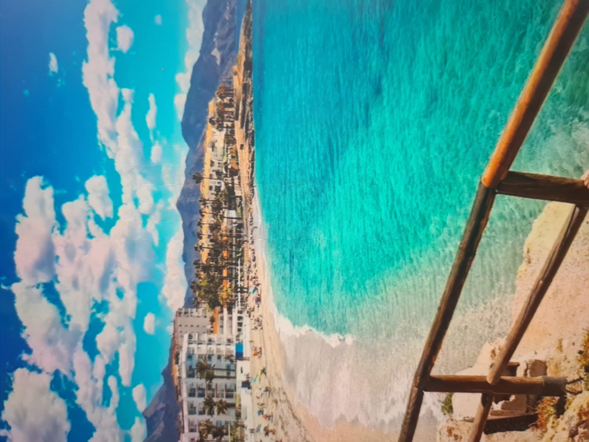 The image displays a vibrant coastal landscape, captured as a photograph of a screen, evidenced by visible pixel grid patterns and slight color banding. The image is currently oriented 90 degrees clockwise from its natural landscape view.
In the foreground, a rustic wooden railing or fence made of logs is visible on the right side, indicating an elevated viewpoint, possibly from a cliff or lookout. Beyond this, a long, sandy beach stretches along the coastline. The clear, bright turquoise water of the ocean laps at the shore with gentle white waves, transitioning from shallow clarity near the beach to a deeper blue further out.
Along the curve of the beach, a dense array of multi-story buildings, likely hotels or apartments, line the shore. These structures are predominantly white or light-colored, featuring numerous balconies. Palm trees are scattered among the buildings, adding to the tropical or subtropical feel of the location. Small figures of people can be observed on the beach, suggesting recreational activities such as sunbathing or swimming.
In the background, rugged mountains rise, forming a natural boundary to the coastal plain. The sky above is a brilliant blue, dotted with scattered white, fluffy clouds. The scene conveys a sunny daytime setting with clear weather. No discernible text is visible within the image.