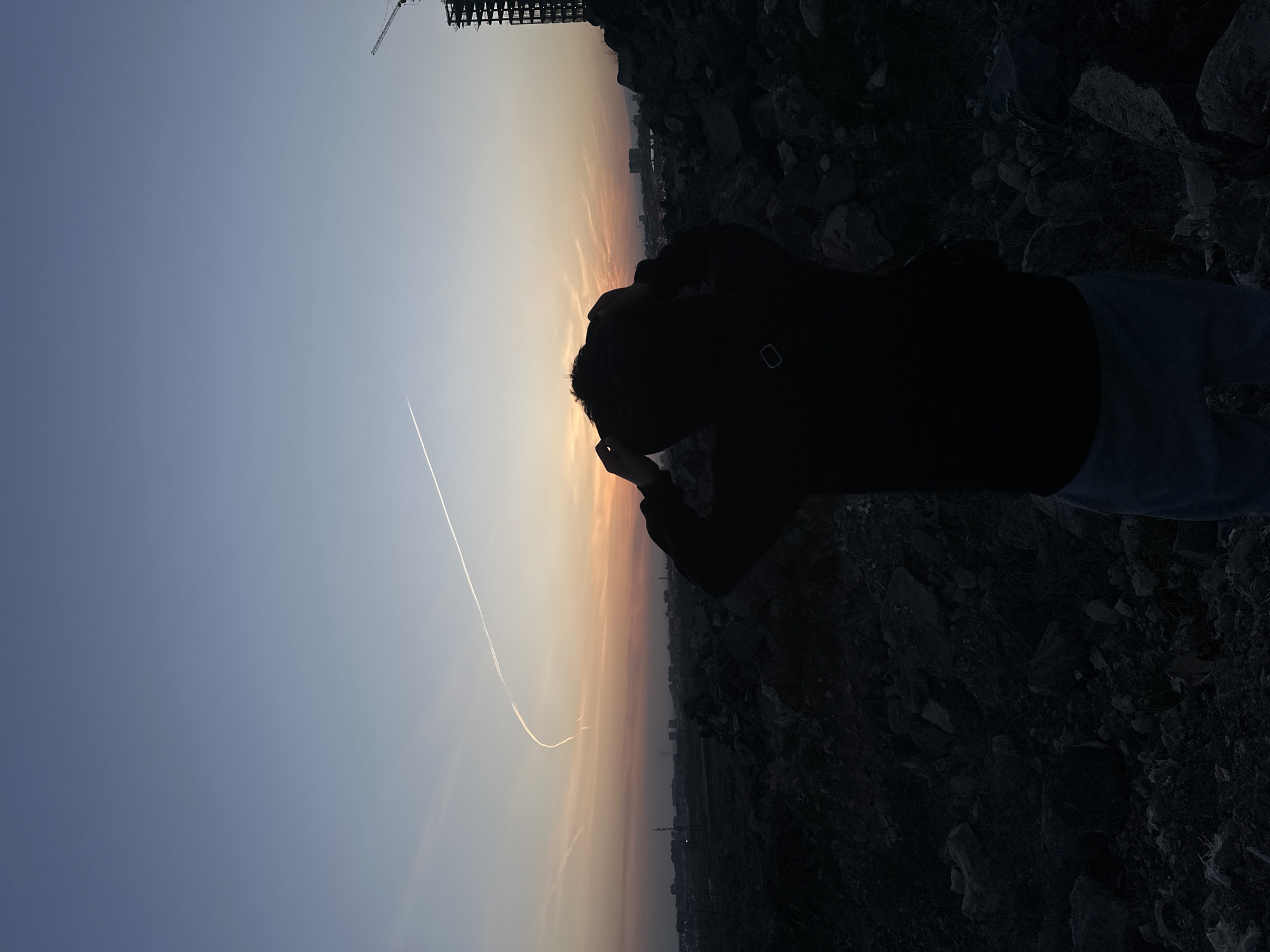 The photograph, taken at a low angle, shows a person silhouetted against a gradient sky transitioning from light blue to orange. A contrail cuts across the sky. The person is wearing a dark jacket, with their hand near their head. They appear to be standing on rocky ground. In the background, there are building silhouettes. A crane is partially visible in the upper left corner. The photo seems to have been taken during sunset or sunrise. The location is an elevated position in an urban area.