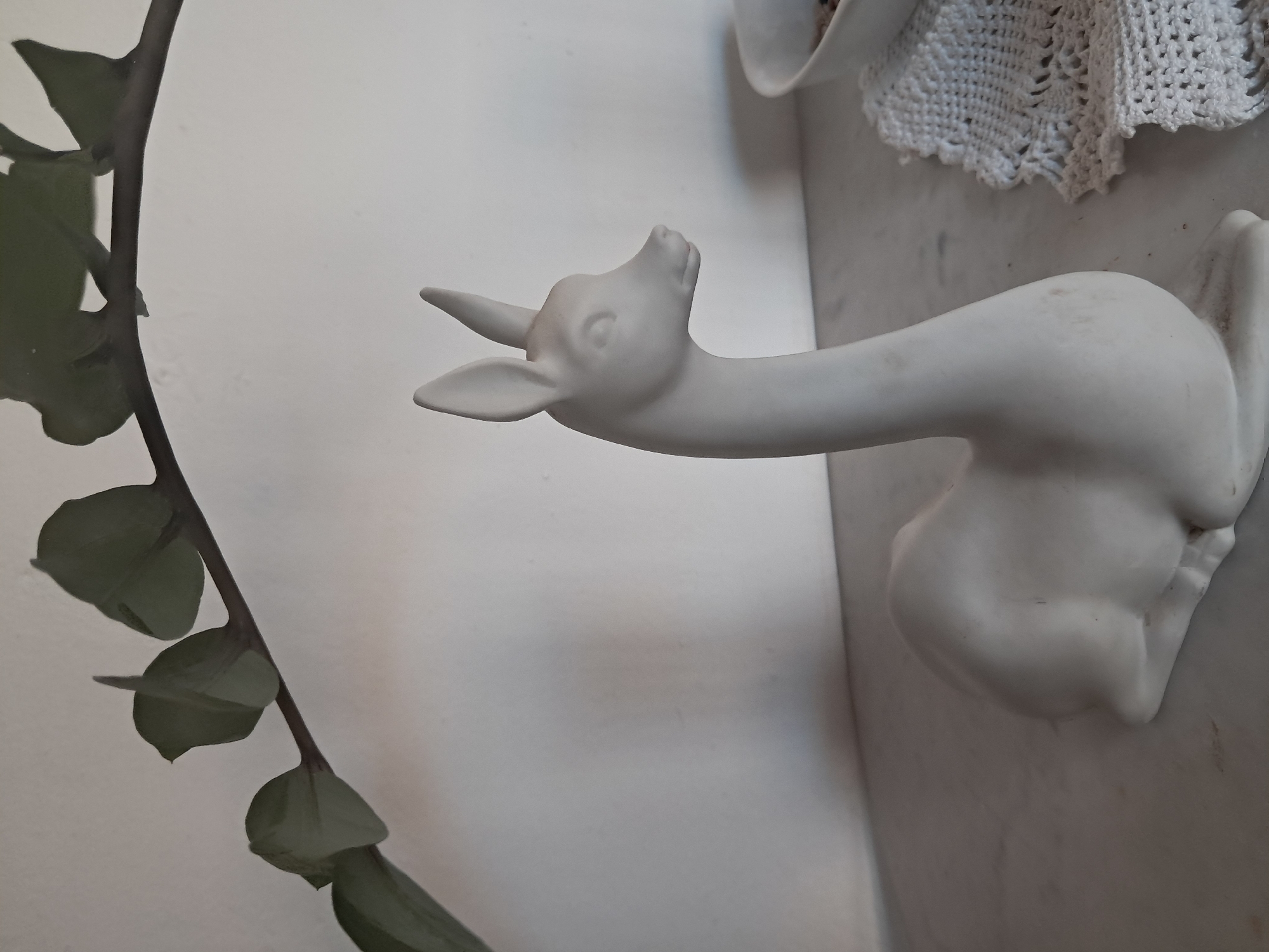 A porcelain deer statue is on a white surface. A plant with dark green leaves is on the left side of the frame. The background is a white wall. A crocheted doily is partially visible in the upper right corner. The location is unknown, and there is no information to suggest the scene is in Juan Griego, Venezuela.