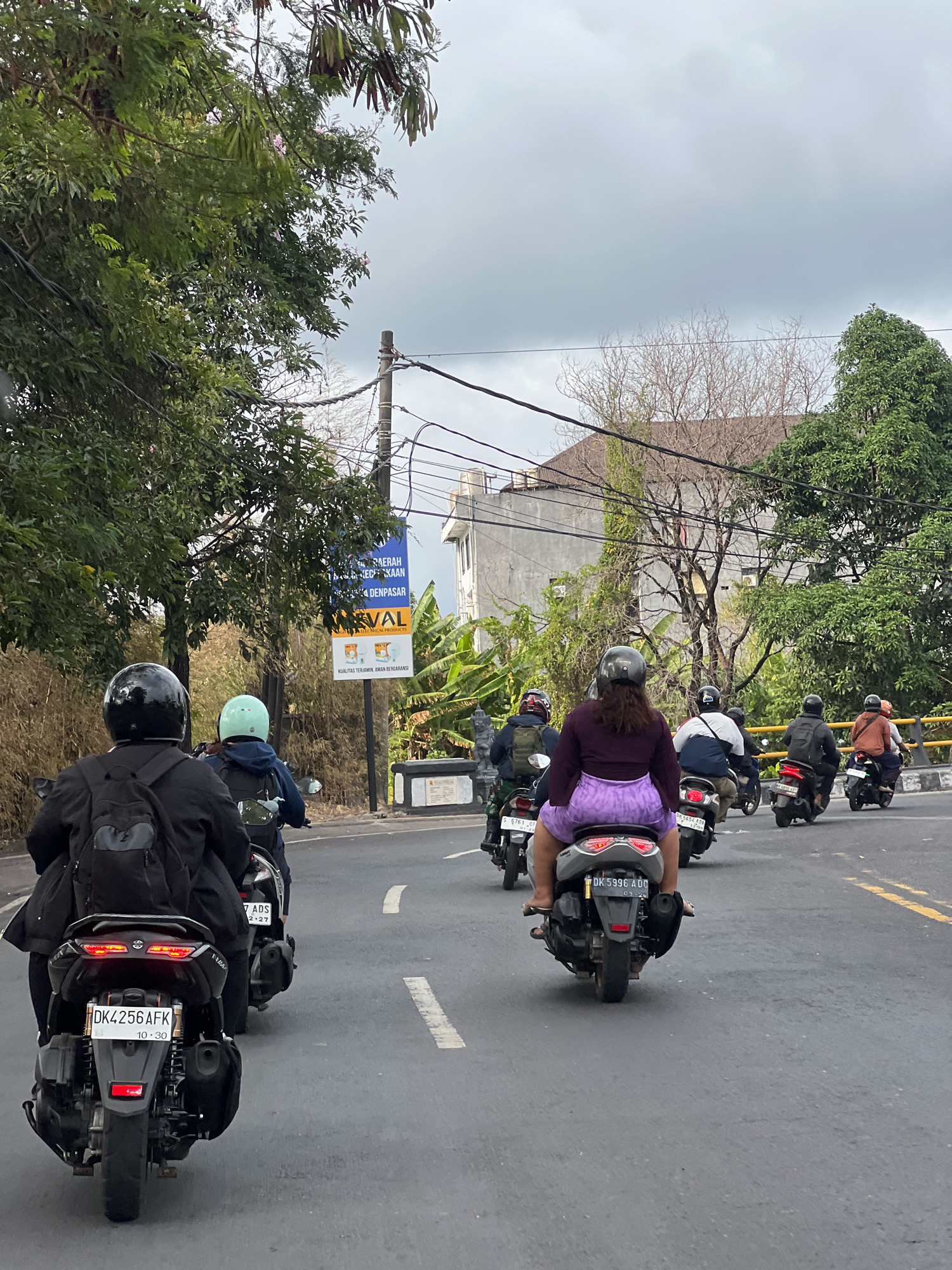 The image depicts a street scene in Denpasar, Indonesia, with multiple individuals riding scooters on a paved road. The perspective is from behind the leading scooter, looking forward. The sky is overcast. Trees line the left side of the road, and buildings are visible in the background. A billboard is also present on the right side of the road, featuring text in Indonesian and an image of medical products. The scooters are of various models and colors, and the riders are wearing helmets. Some riders have backpacks. The road has white and yellow lane markings. The overall scene suggests a typical urban environment in Denpasar, with motor vehicles being the primary mode of transportation.